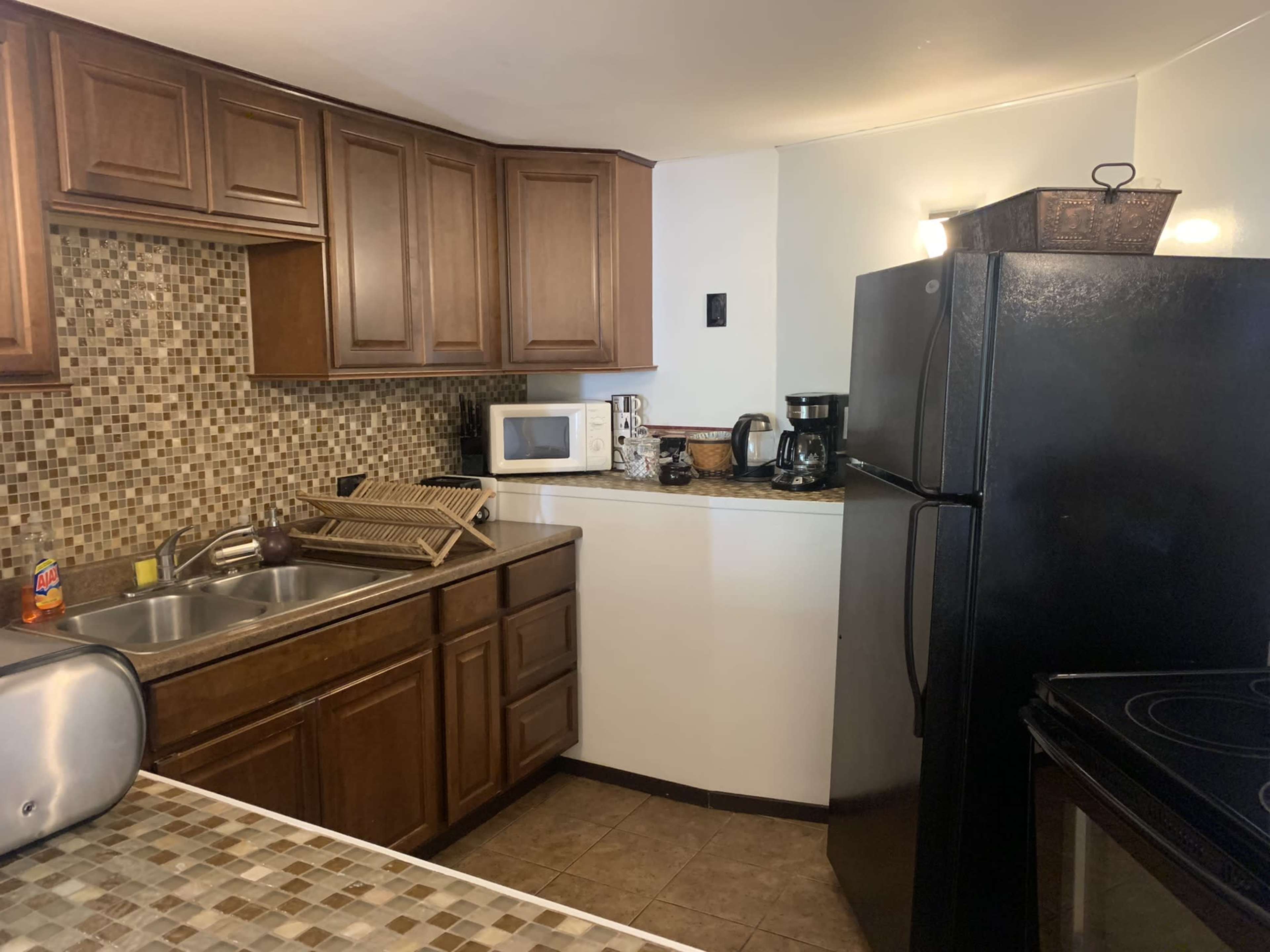 A compact kitchen featuring dark wood cabinets, a black refrigerator, a microwave, a coffee maker, and a tile backsplash.