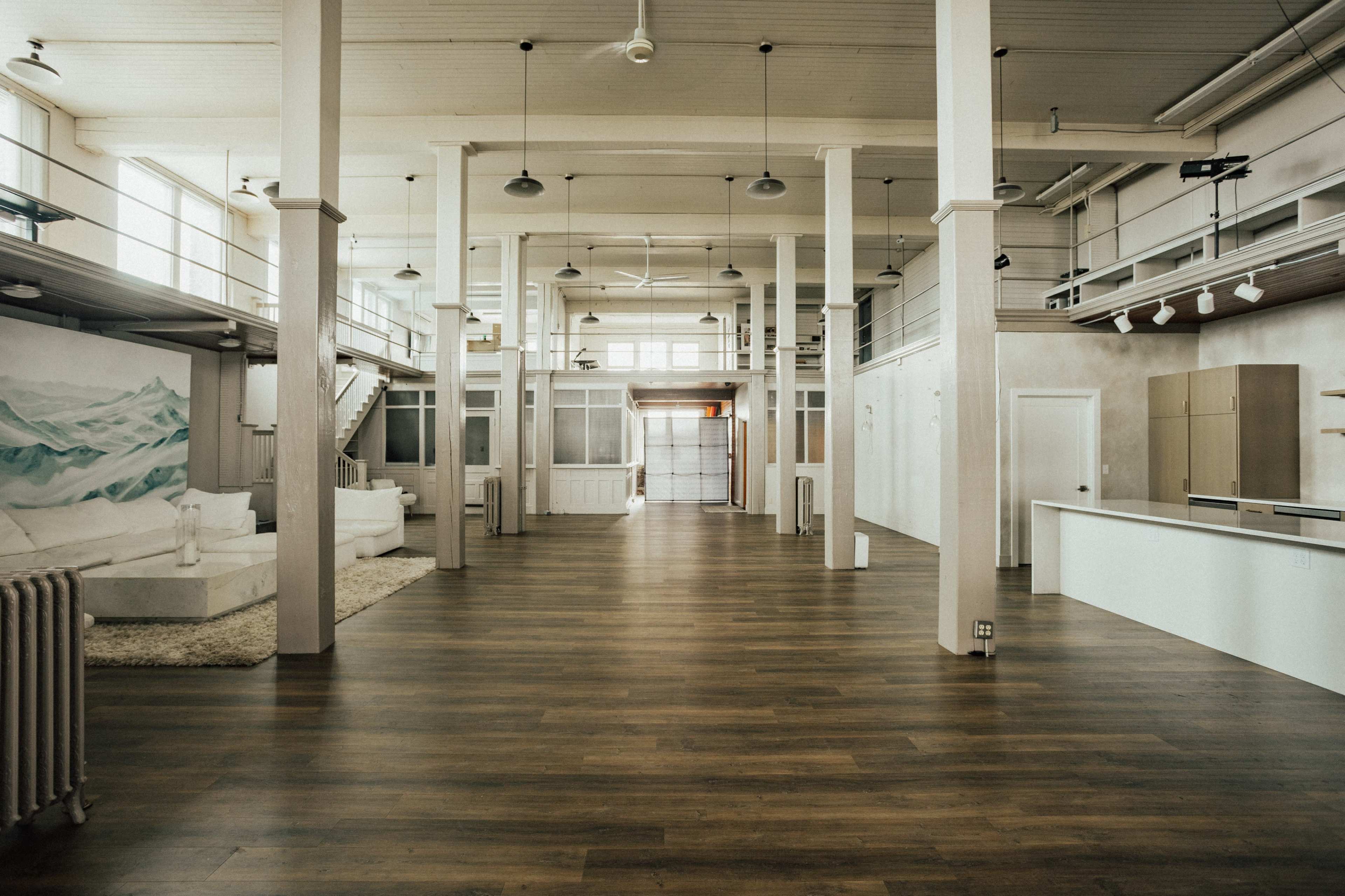 A spacious, modern interior with wooden floors, tall white columns, and large windows, featuring minimal furnishings and an open layout.