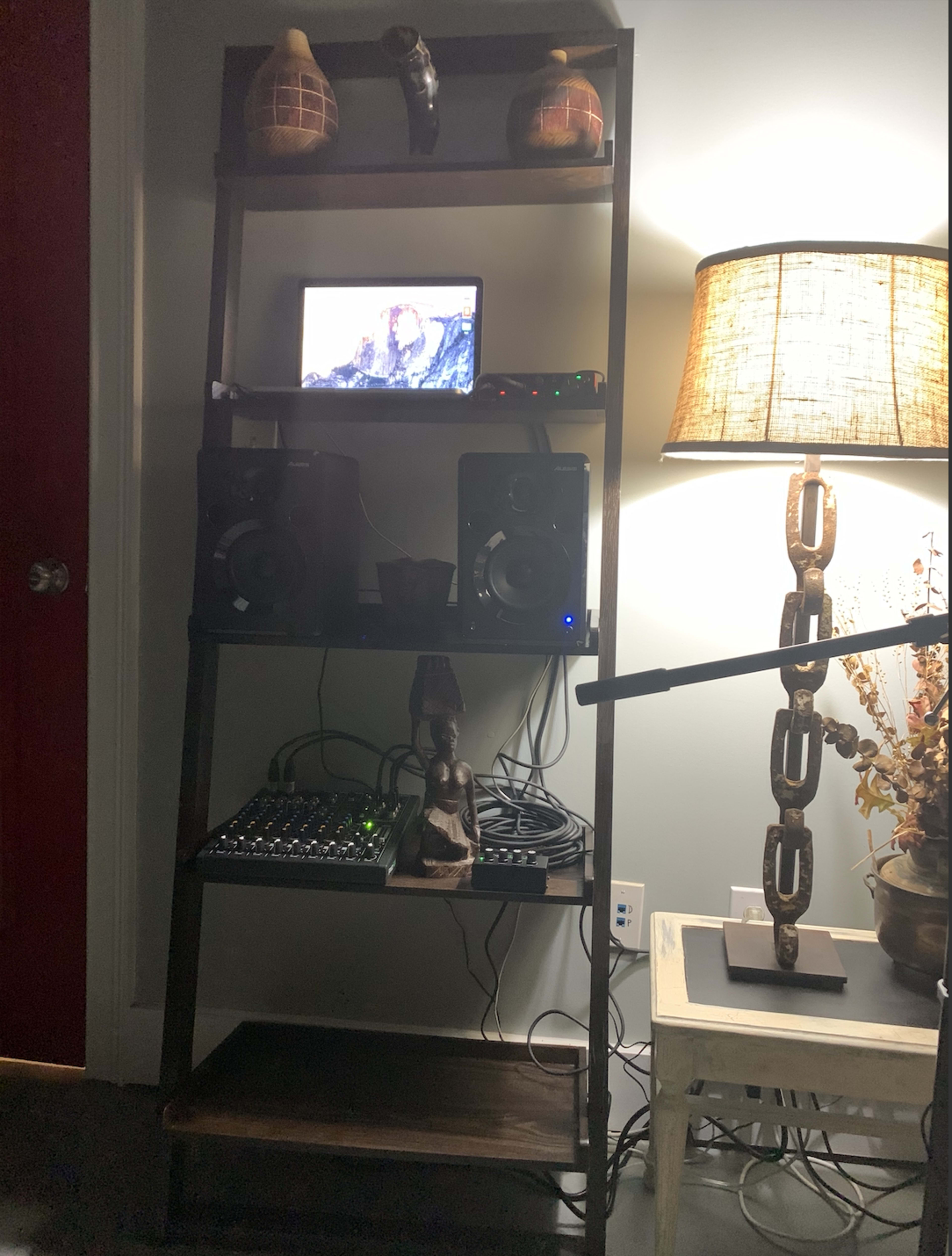 A wooden shelf holds a monitor, speakers, a mixer, and decorative items, with a lamp and a piece of furniture nearby.