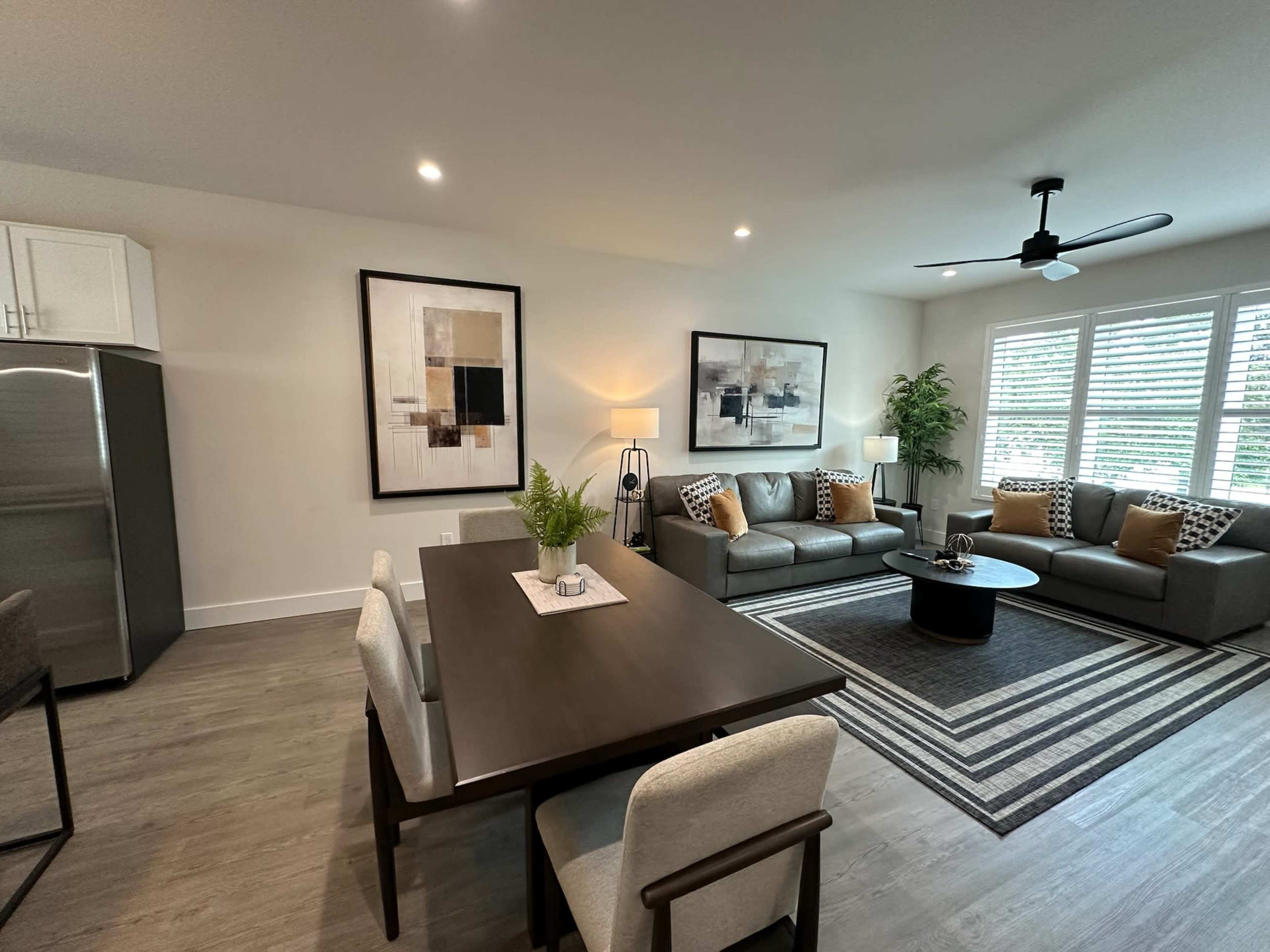 A modern living room and dining area featuring a gray sofa set, a round coffee table, and a dining table with chairs, all set against a backdrop of large windows with blinds.