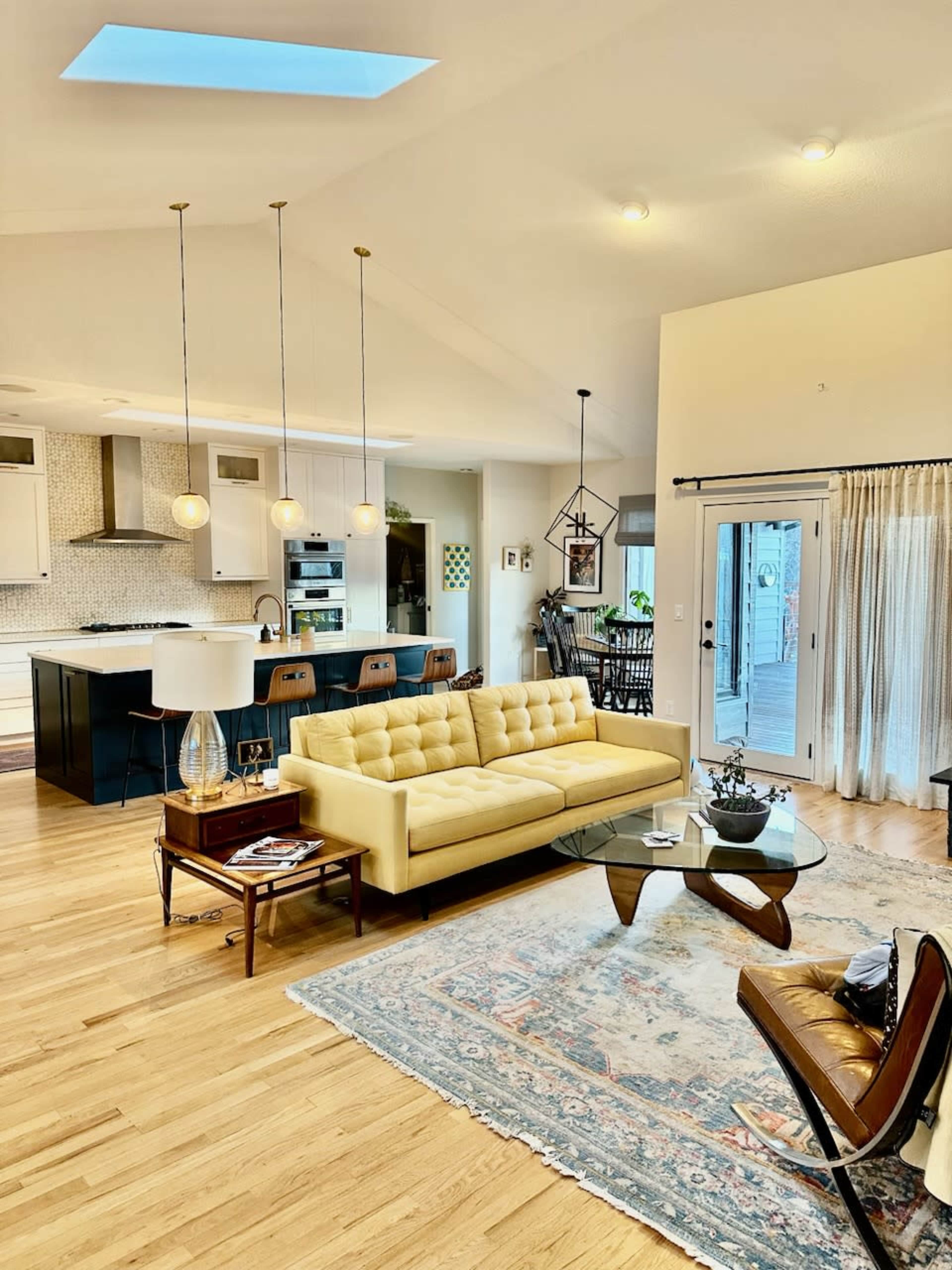 A bright living area features a yellow tufted sofa, a glass coffee table, and a modern kitchen visible in the background.