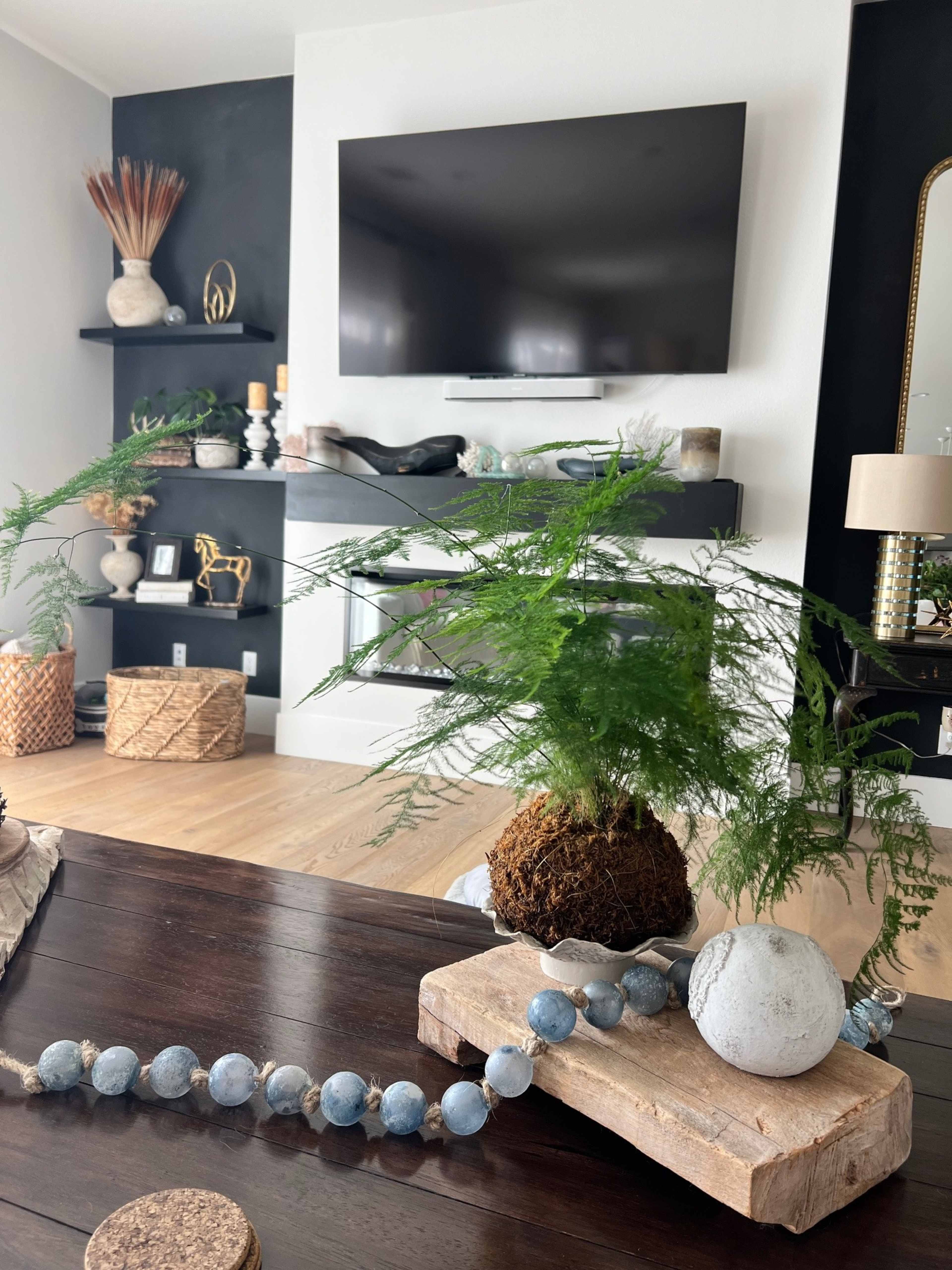 The image shows a modern living room featuring a mounted television, a decorative shelf with various items, and a table adorned with a plant arrangement and a beaded decor piece.
