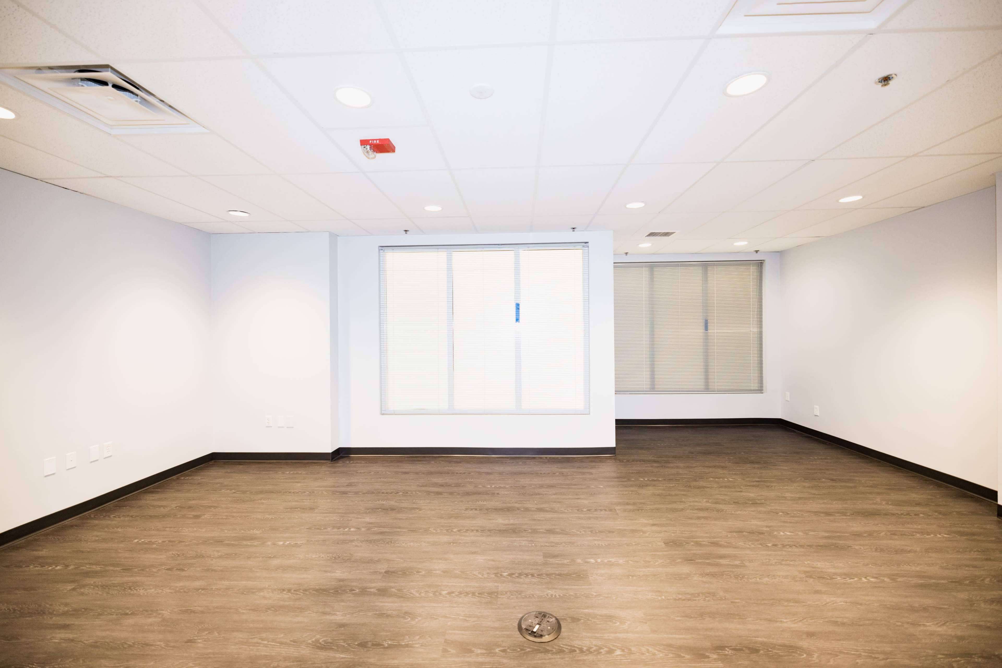 The image shows an empty room with light-colored walls, a wooden floor, and a window with blinds.