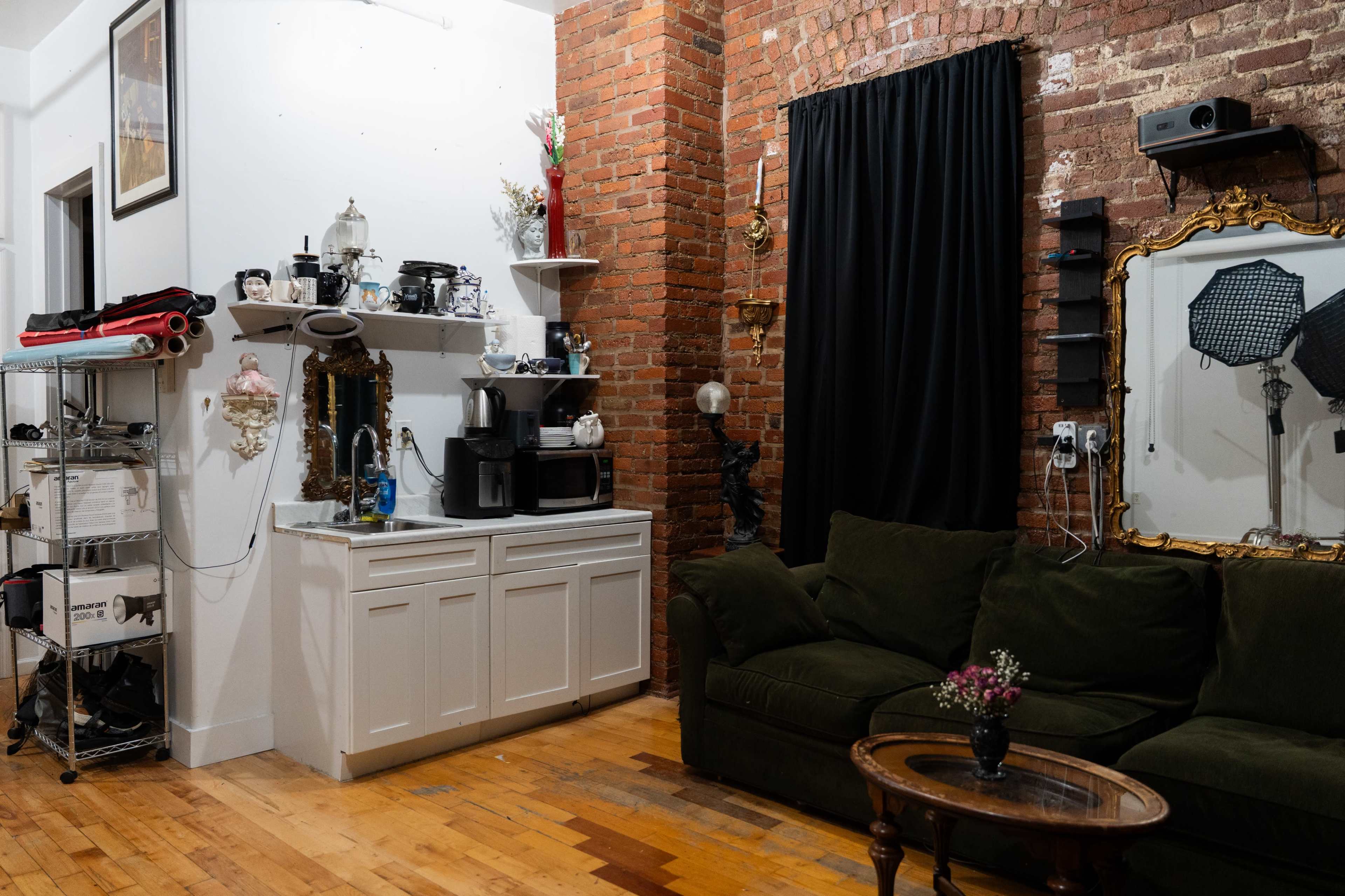 The image depicts a cozy room with exposed brick walls, a dark curtain, a small kitchen area with appliances, and a green couch facing an ornate mirror with various decorative items.