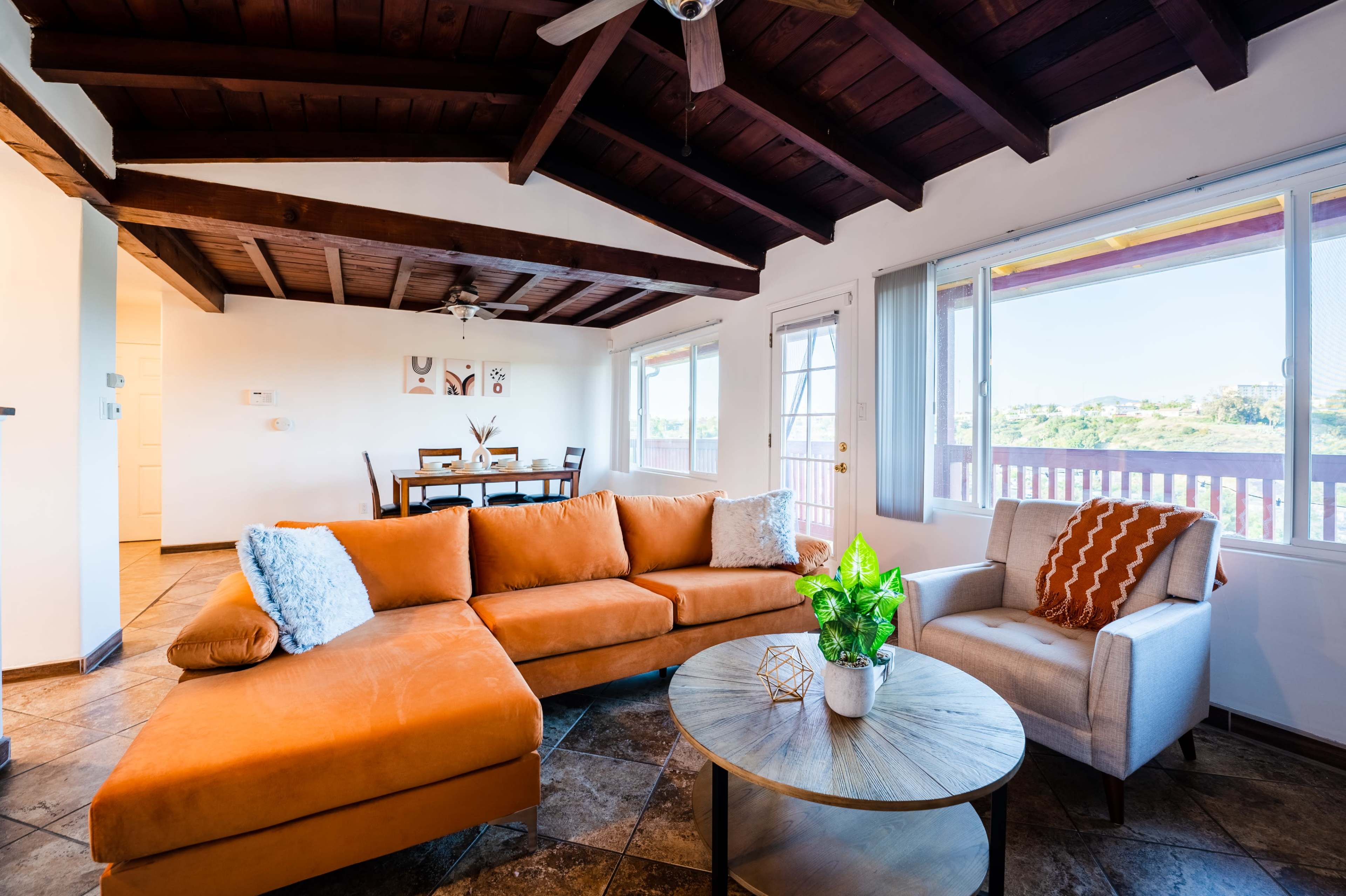 The image shows a cozy living room featuring an orange sectional sofa, a round coffee table, and large windows with views of the outdoors.