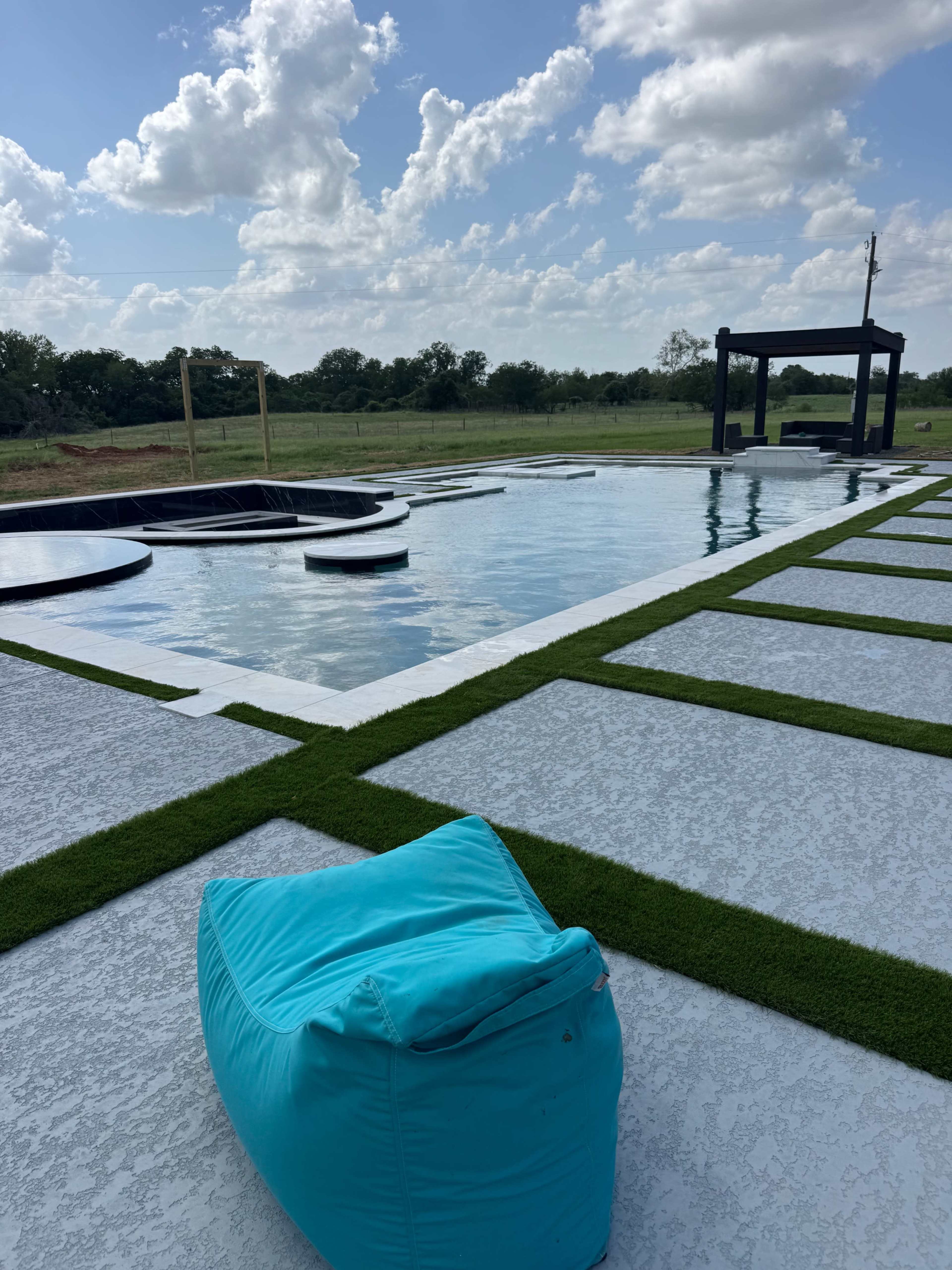A blue bean bag chair is positioned on a concrete deck beside a large swimming pool surrounded by grass and trees under a partly cloudy sky.