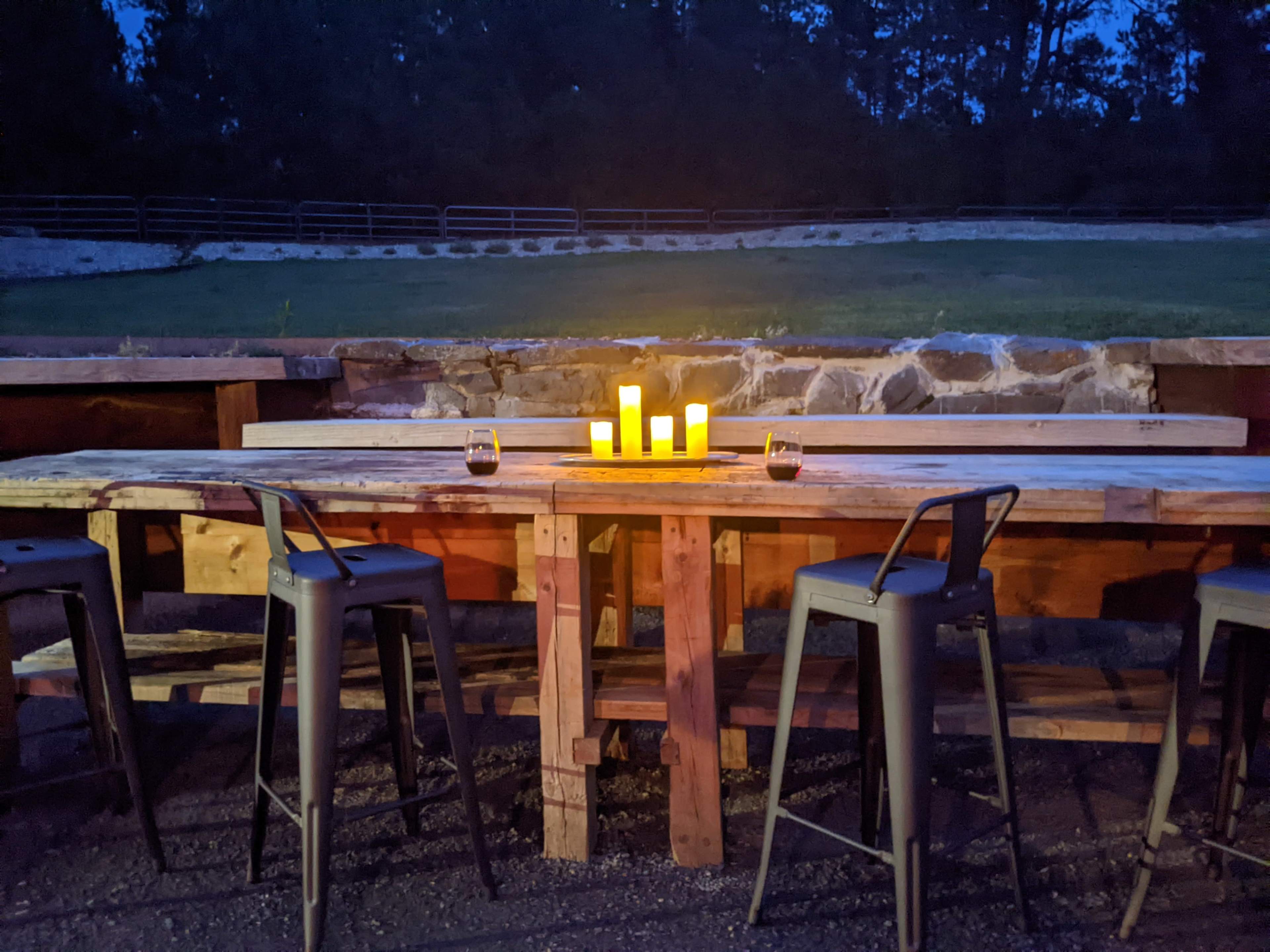 A wooden bar table with high stools is illuminated by candles in a dimly lit outdoor setting.
