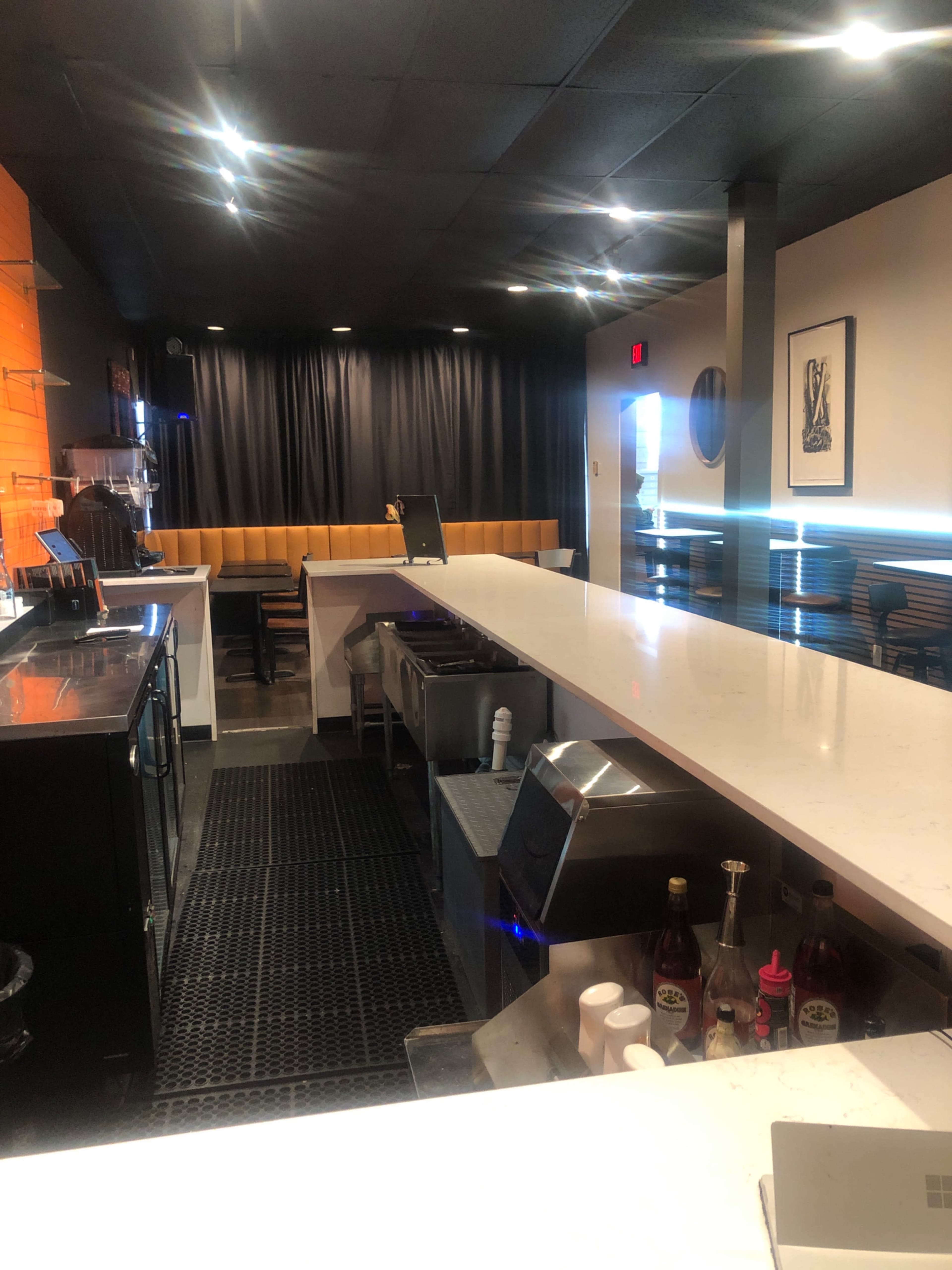 The image depicts a clean, modern bar area with a long countertop, a dark mat, and a backdrop of black curtains.