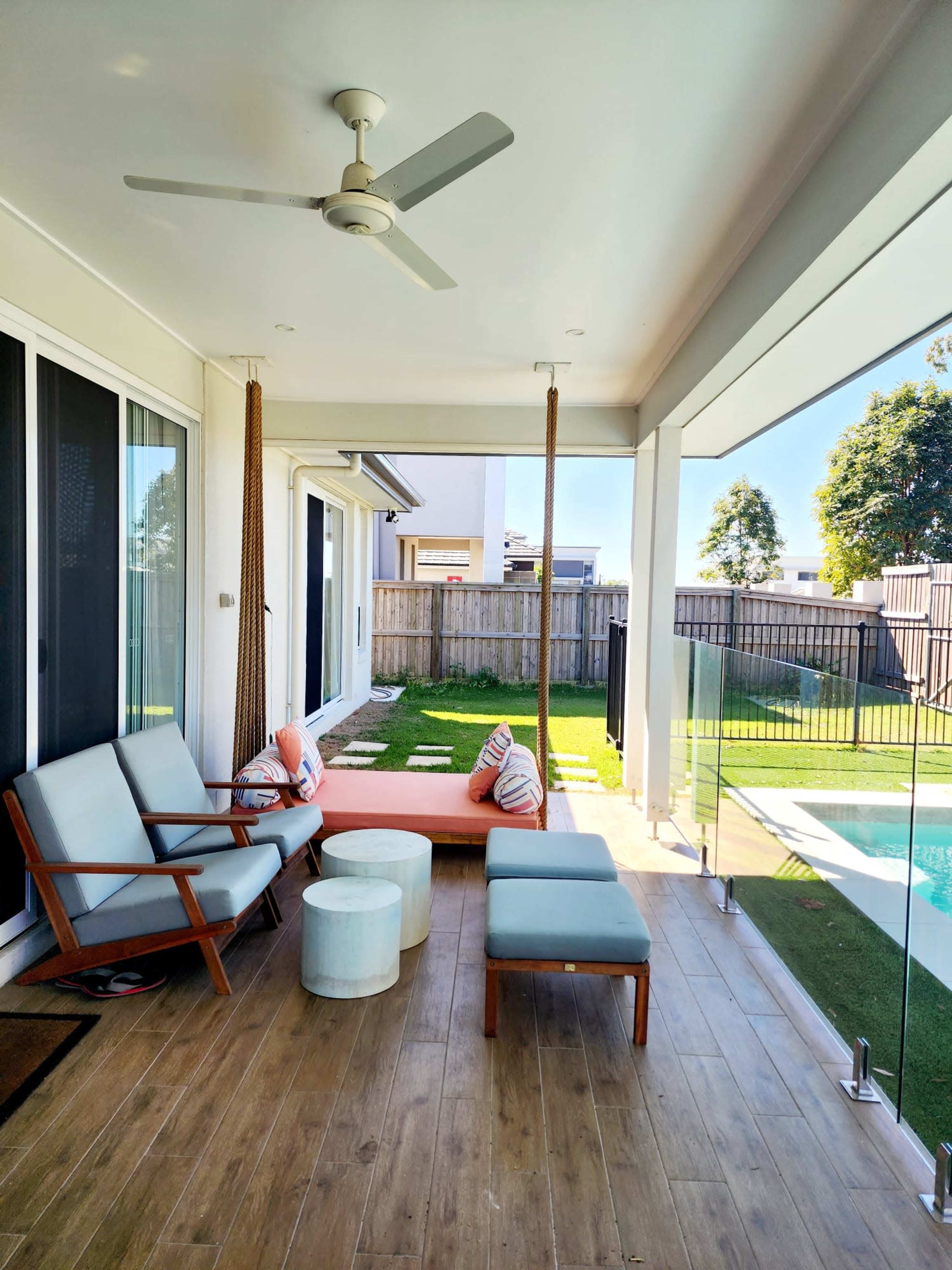 The scene shows a well-lit patio area with a ceiling fan, featuring two chairs, a swing seat, and a small round table beside a swimming pool.