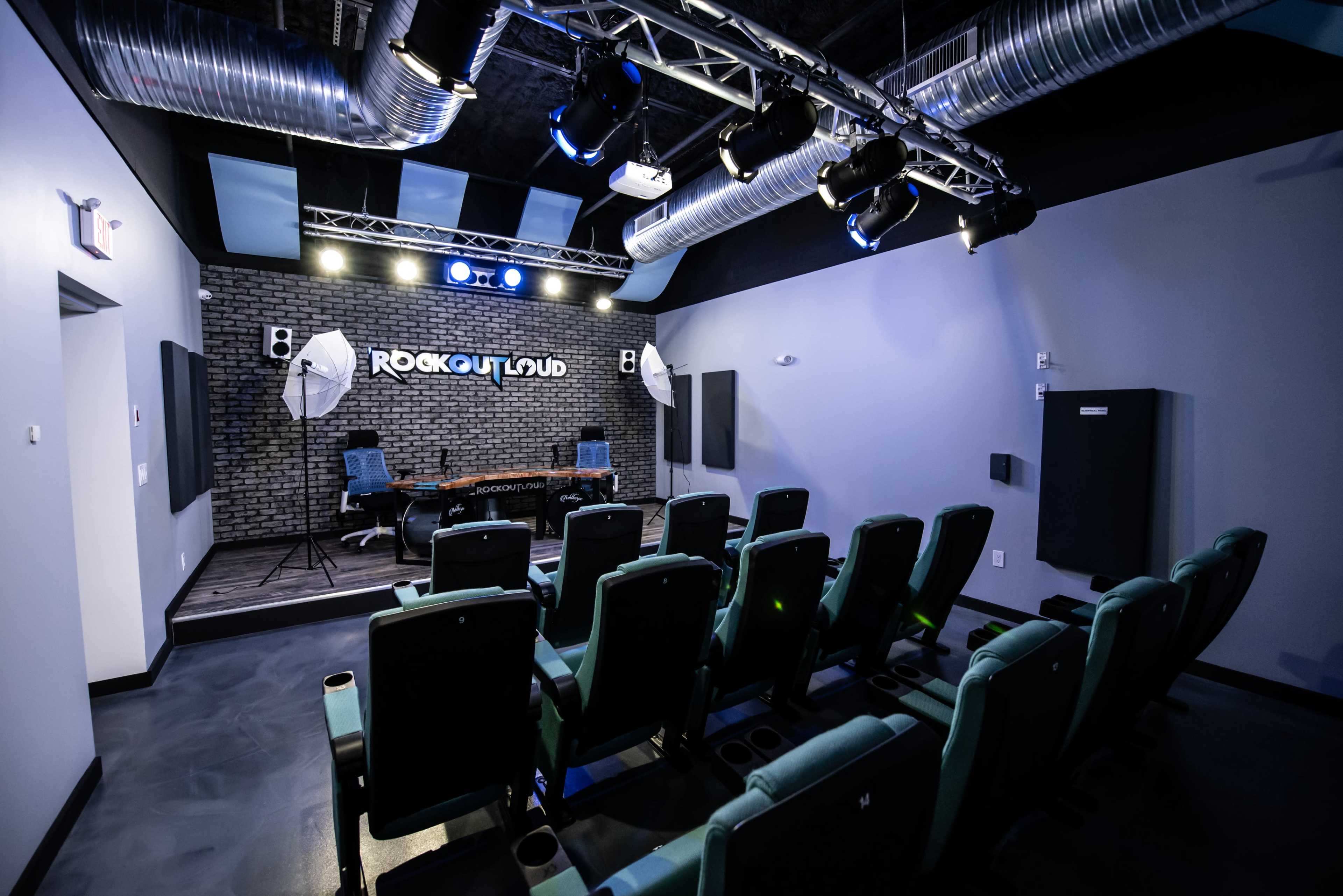 A small performance space with rows of teal chairs facing a stage equipped with lighting and sound equipment.