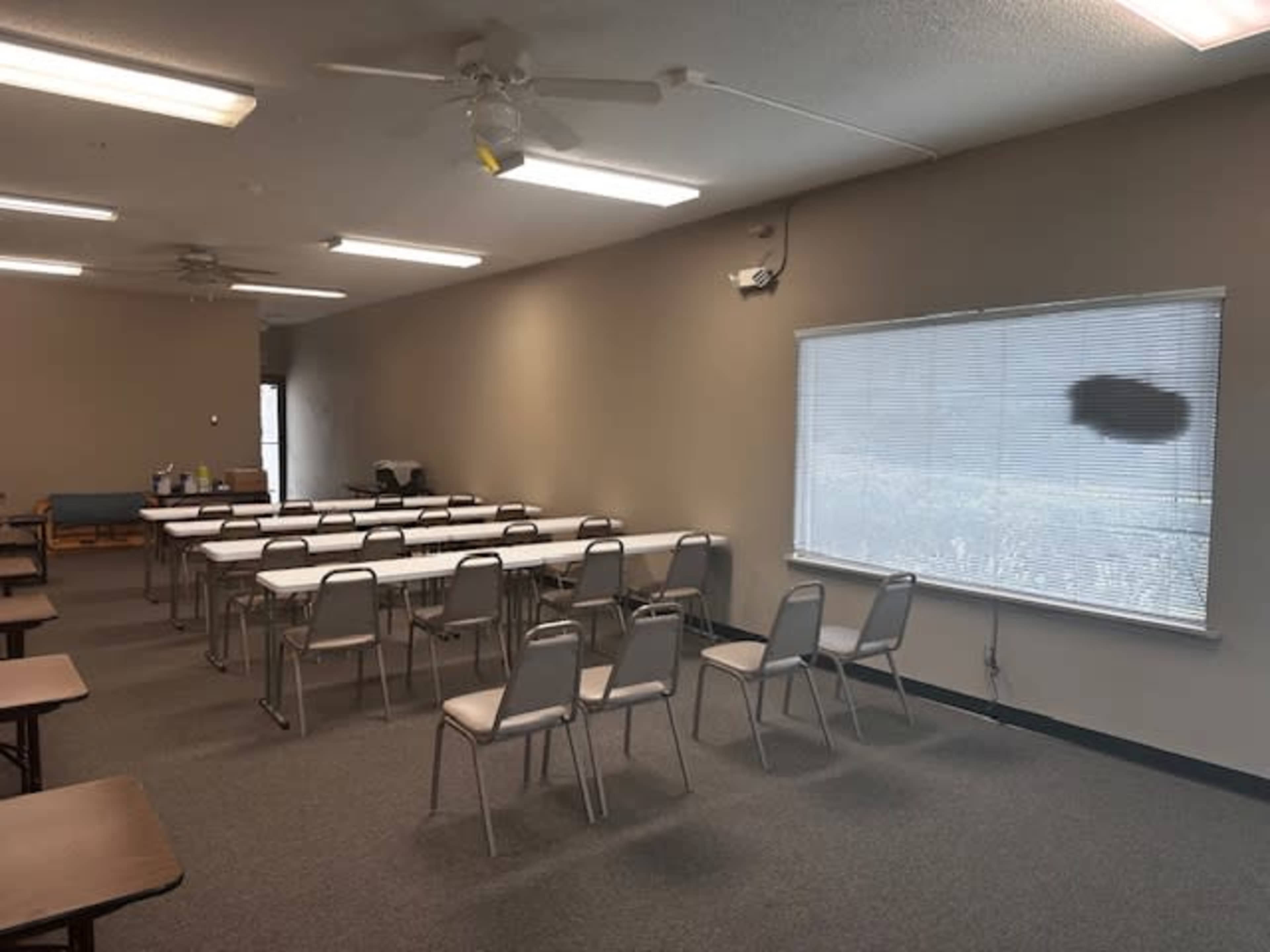A classroom with several rows of tables and chairs arranged neatly, featuring a window with blinds and a plain wall.