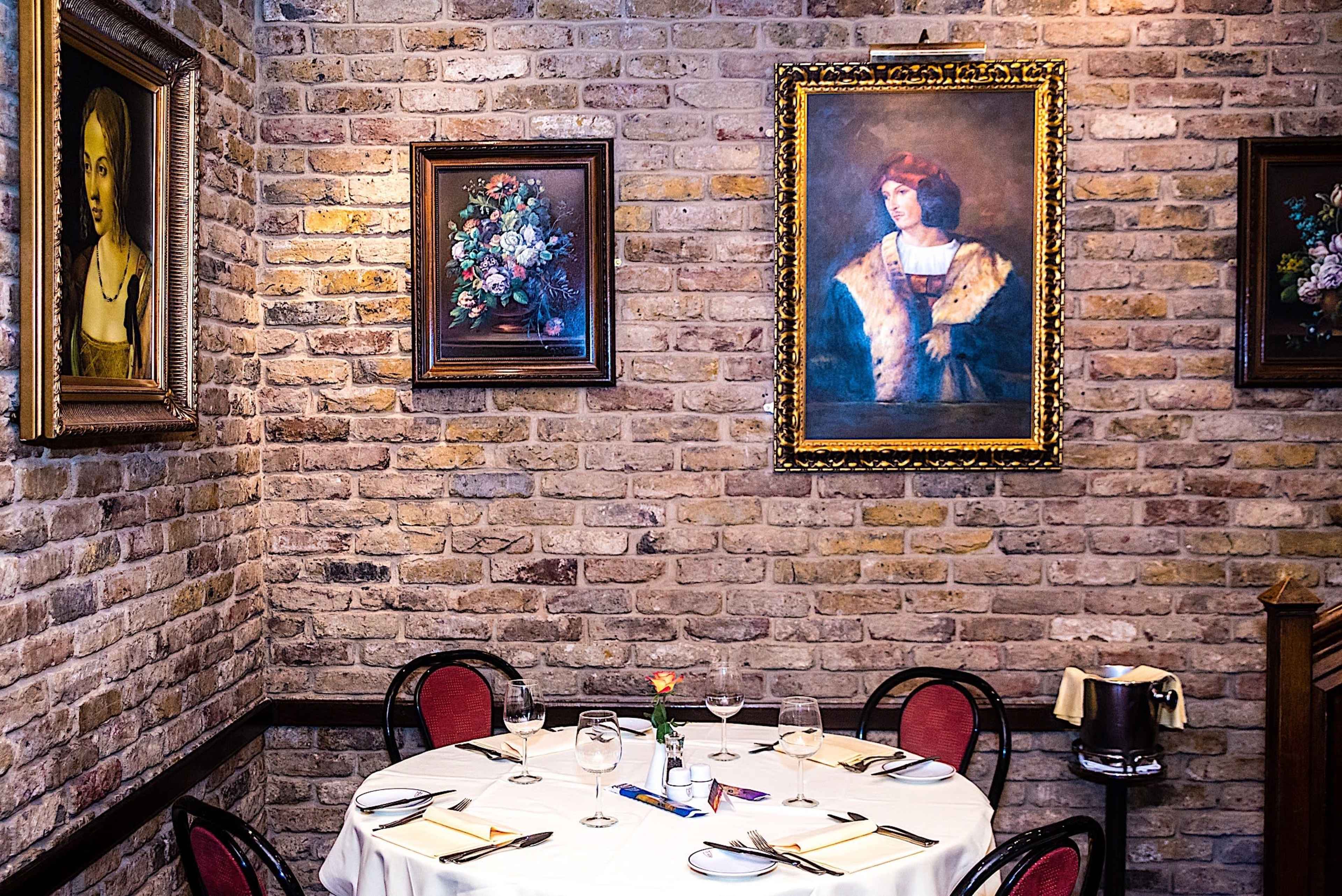 A small restaurant table set for dining in a corner with brick walls adorned with framed paintings.