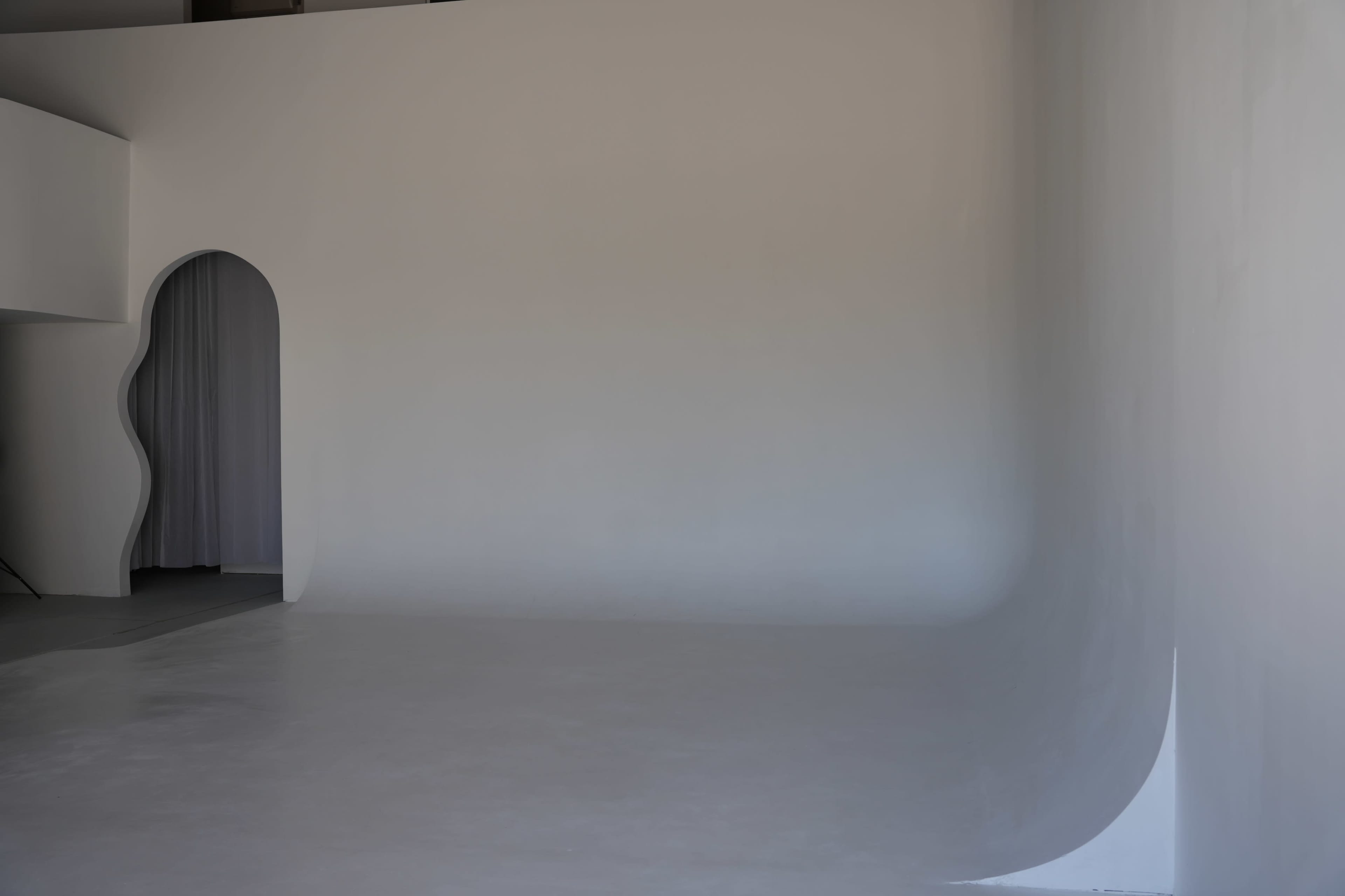The image shows a minimalist interior with a curved white wall and an arched entryway leading to a draped curtain.