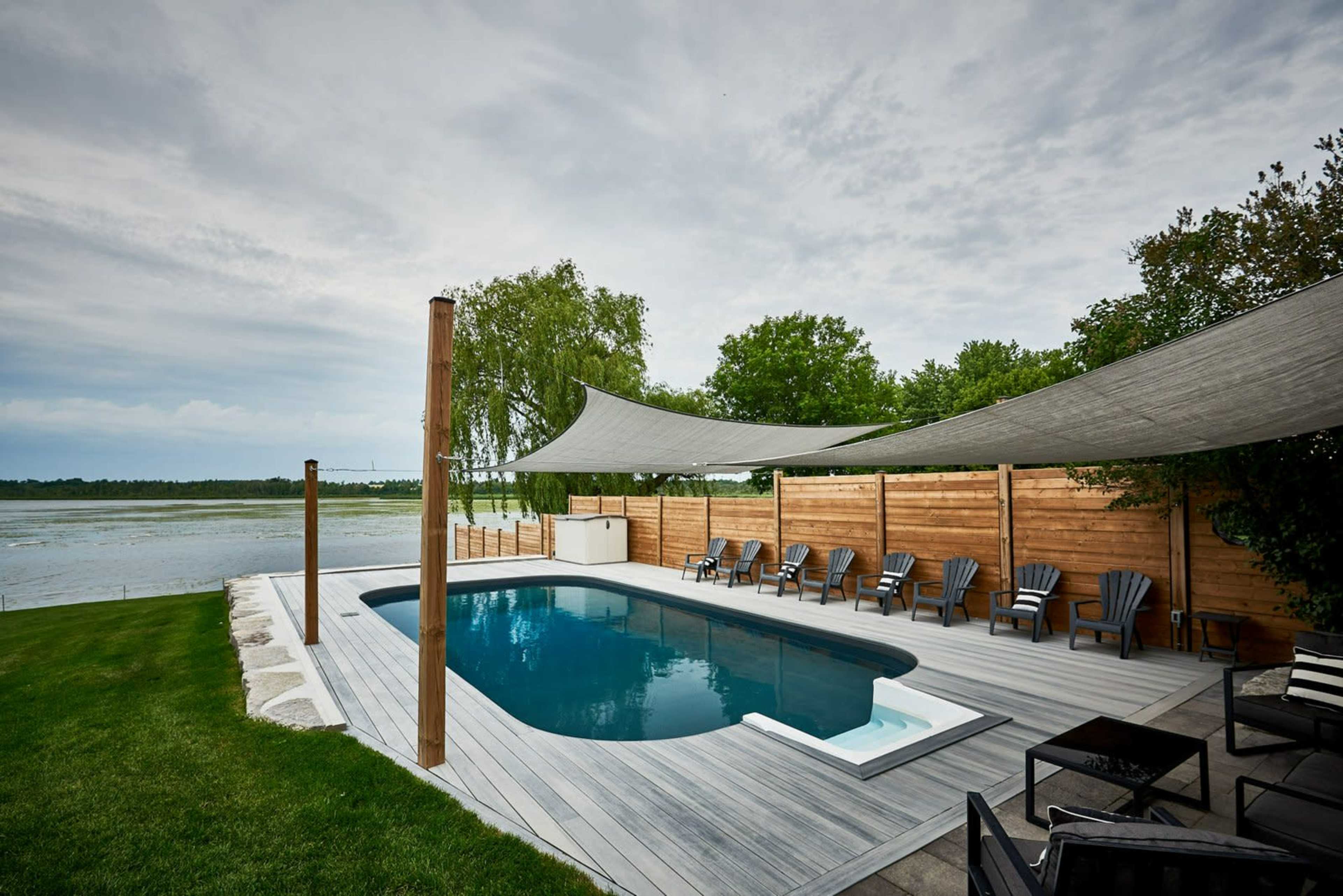 A swimming pool surrounded by wooden decking and chairs, with shade sails overhead, next to a calm body of water and green trees.