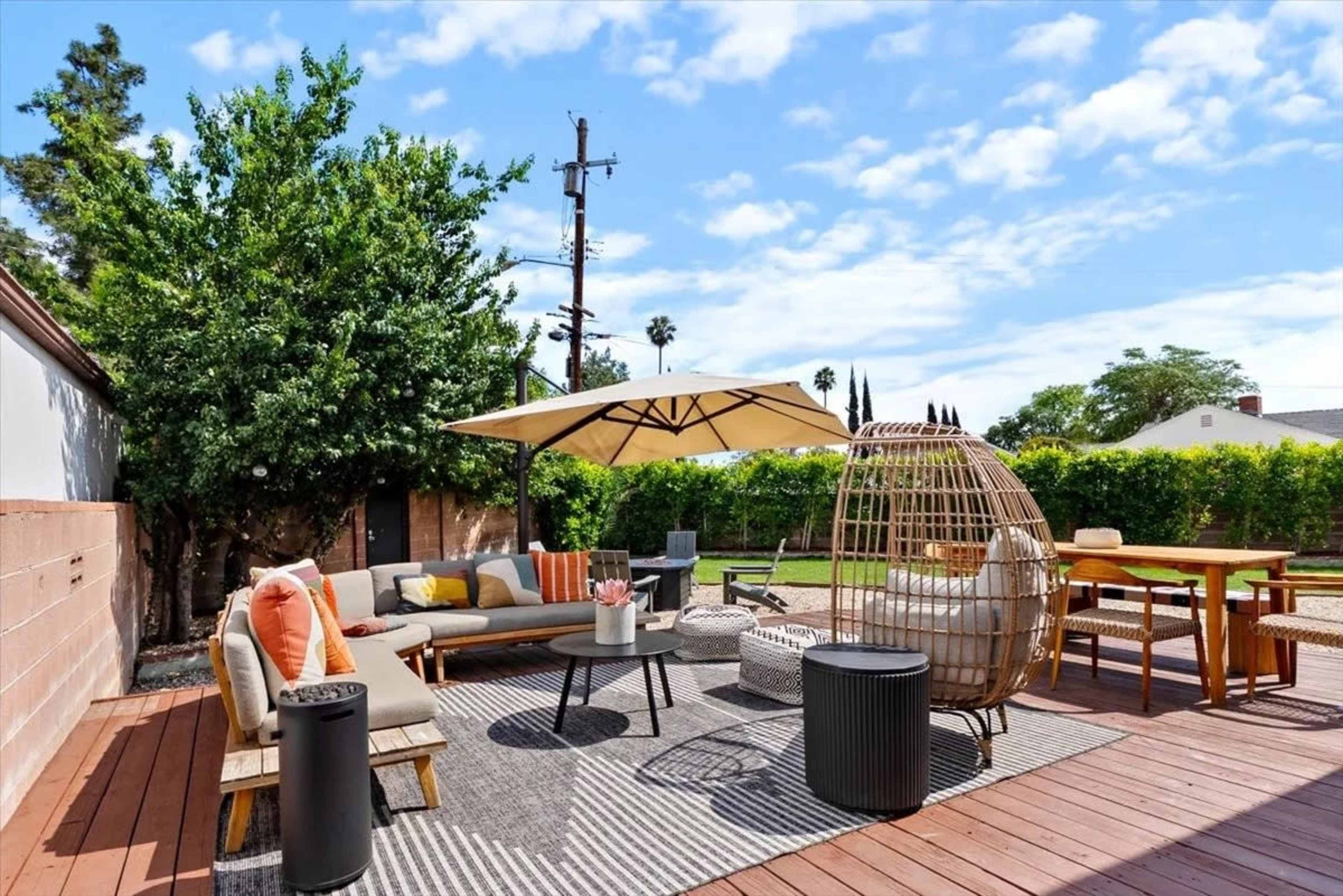 The image shows a wooden deck with patio furniture, including a sofa, a round table, and an umbrella, surrounded by greenery and a clear blue sky.