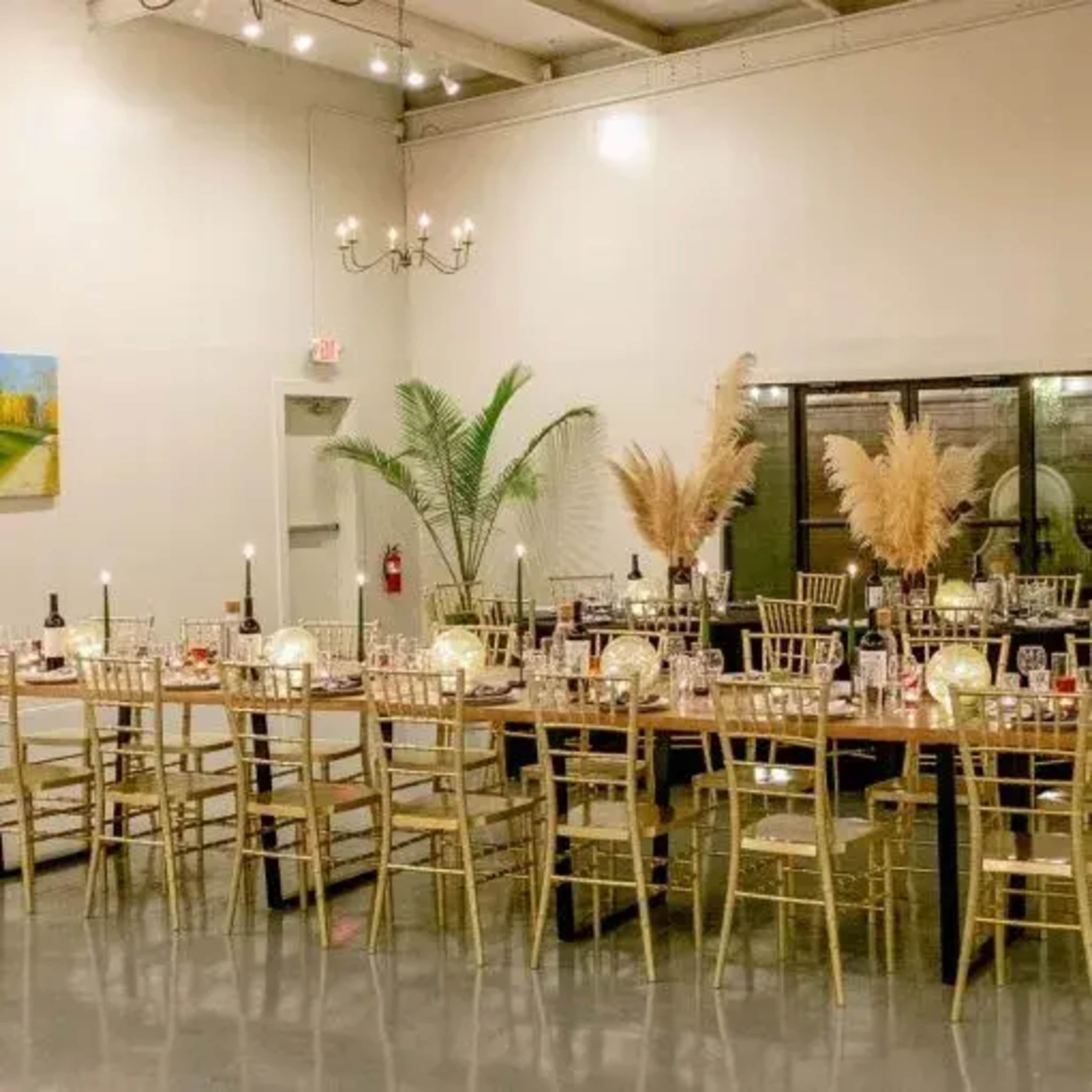 A dining area is arranged with long tables set for a formal event, featuring decorative candles, wine bottles, and plants.