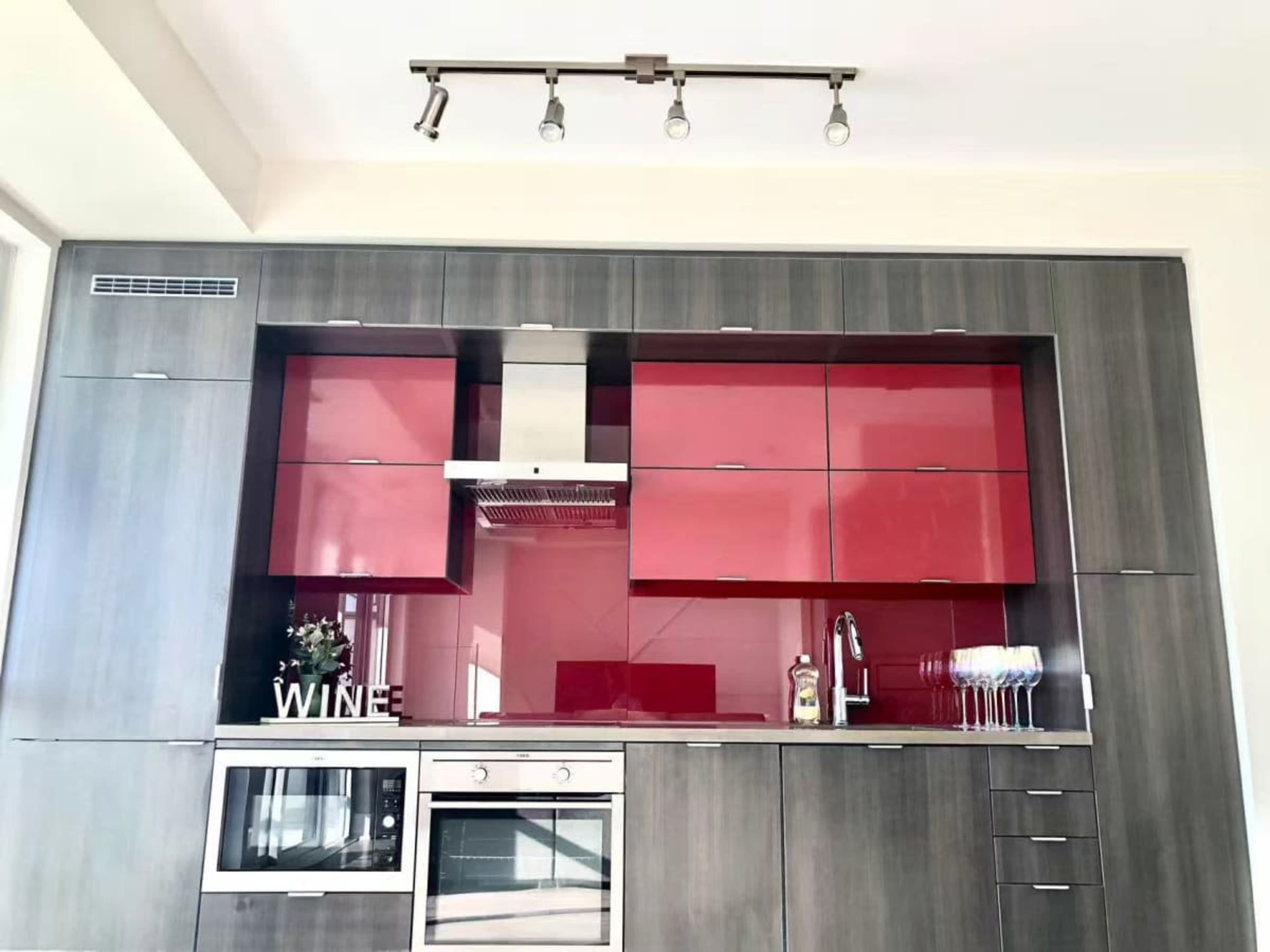 The image shows a modern kitchen with gray cabinetry featuring red accent doors, a stainless steel oven, and a wine display.
