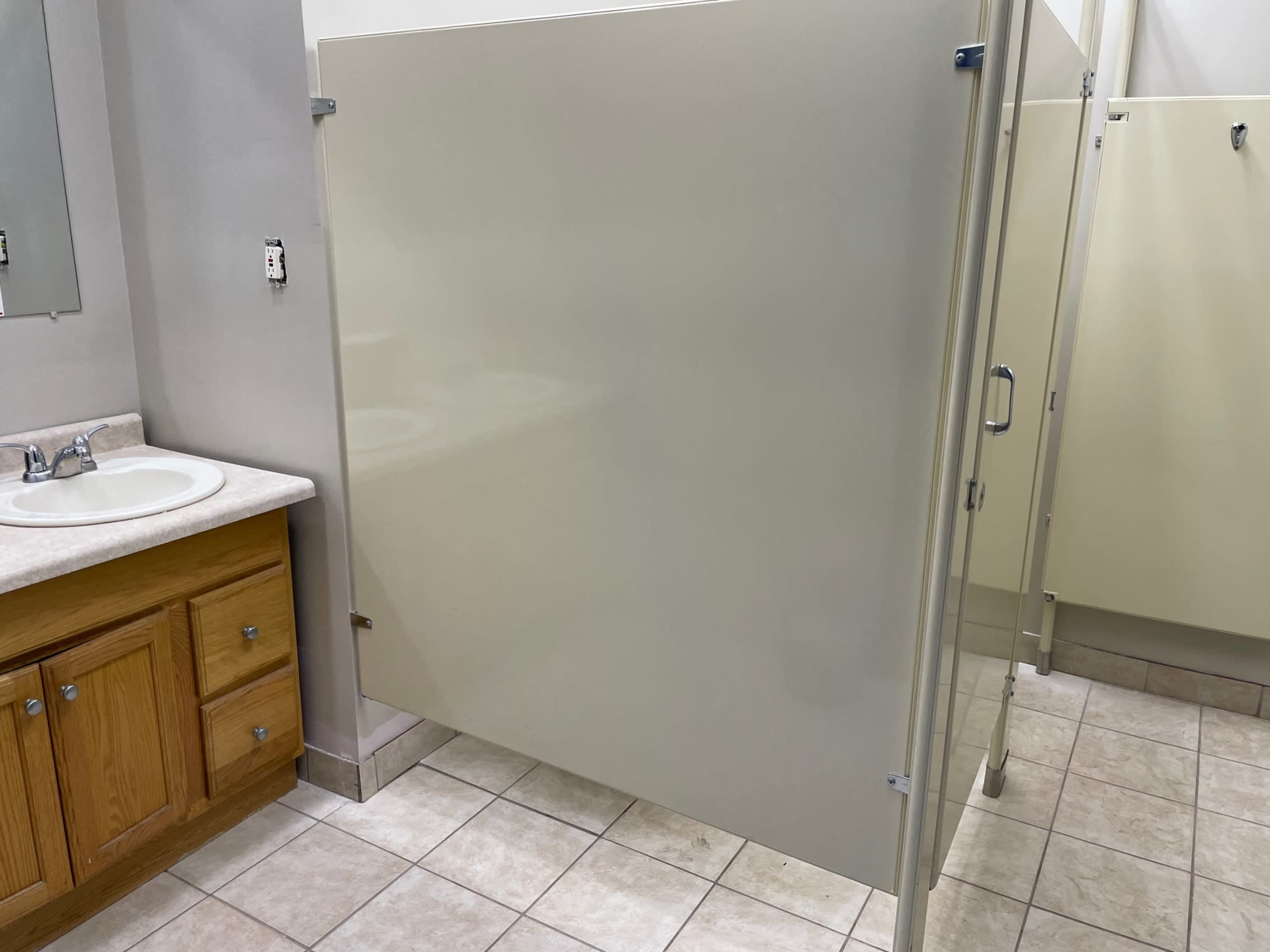 A restroom with a sink, wooden cabinet, and partially enclosed toilet stalls in a tiled space.