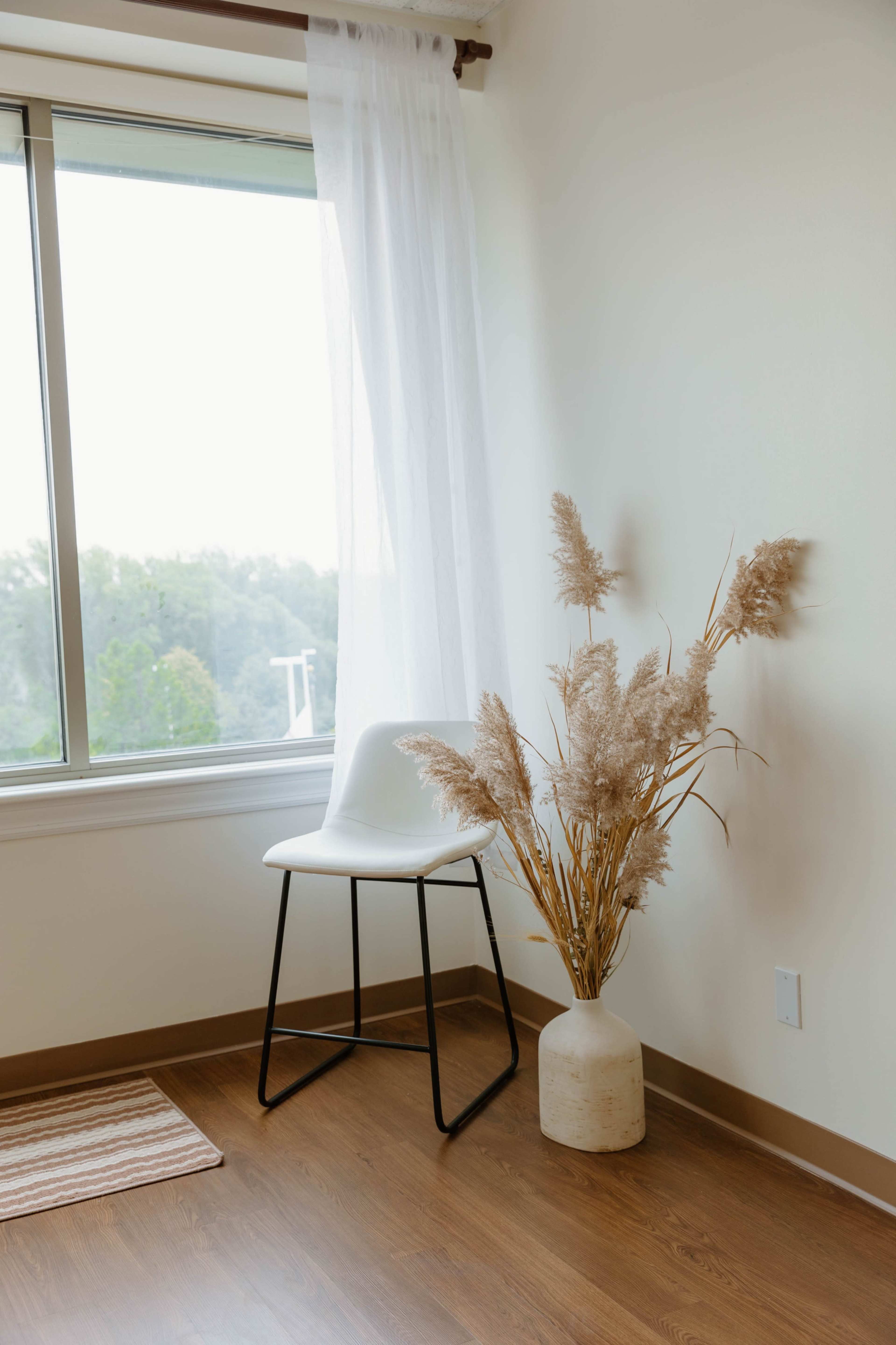 A white chair stands beside a large window adorned with sheer curtains, next to a vase filled with dried plants on the wooden floor.