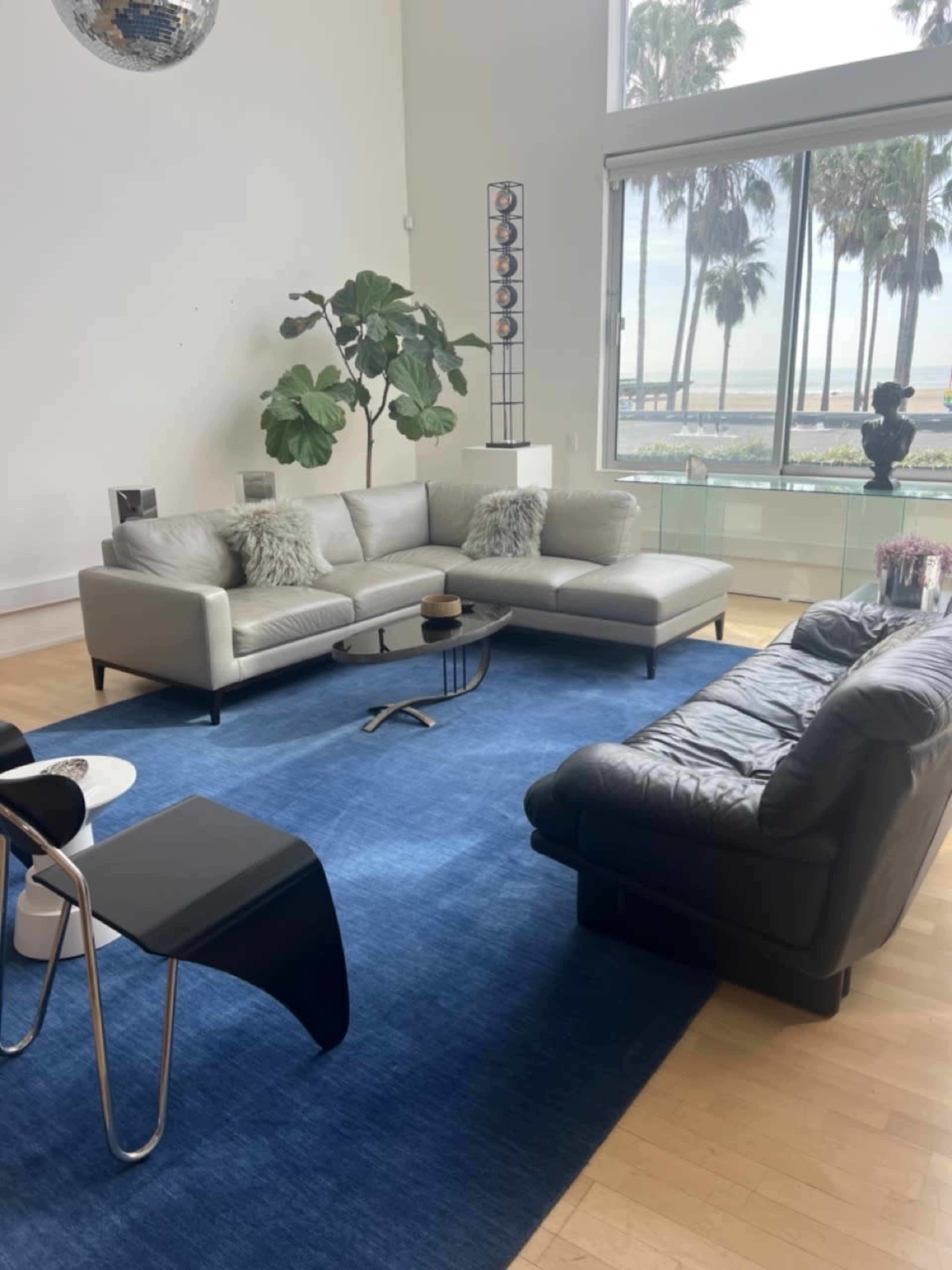A modern living room with a gray sectional sofa, a black leather sofa, a blue area rug, and large windows overlooking palm trees and a beach.