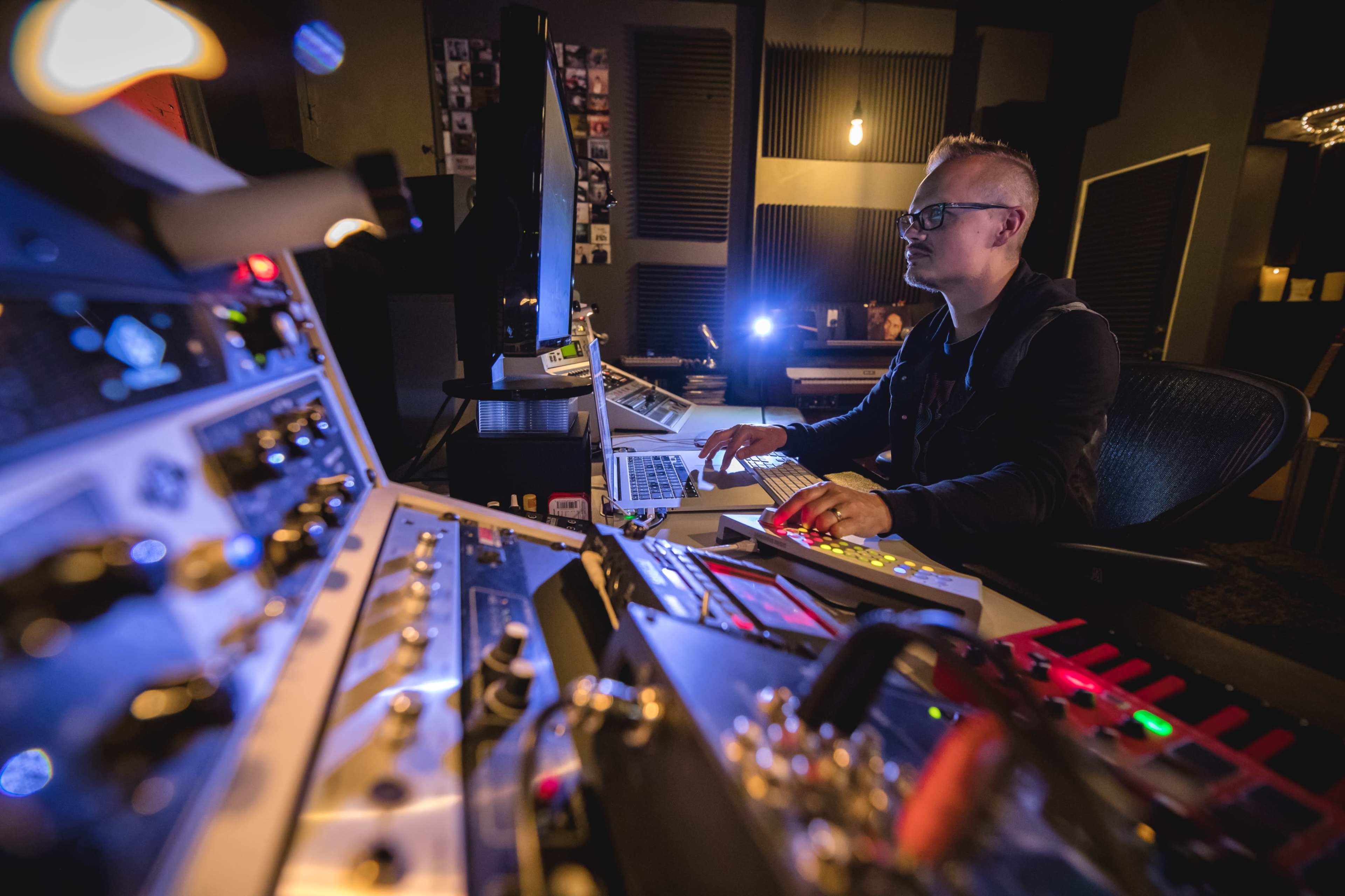 A person works at a music production console surrounded by audio equipment and a computer in a recording studio.