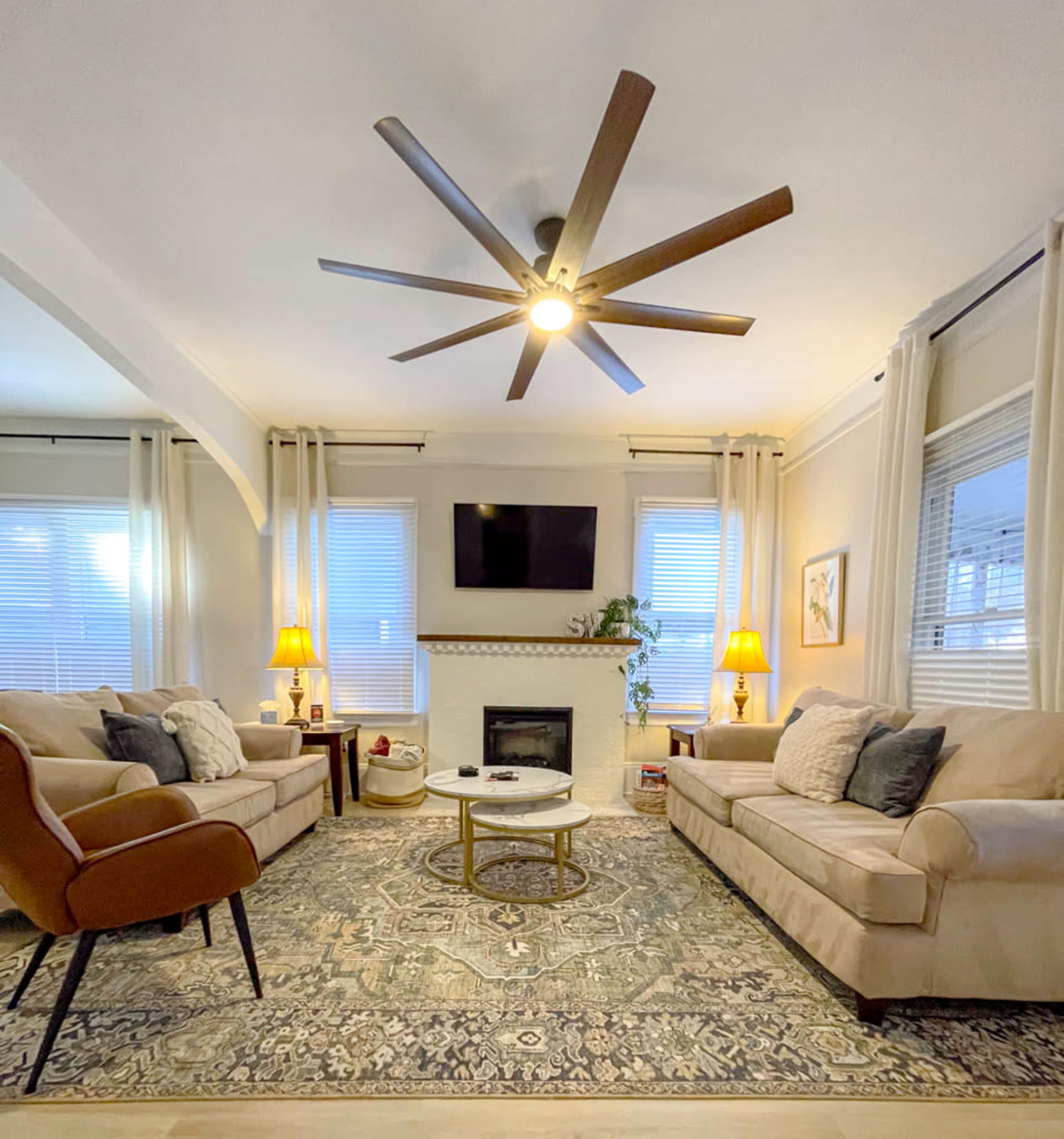 The image shows a cozy living room featuring two beige sofas, a circular coffee table, a fireplace, and a large ceiling fan, all complemented by large windows with curtains and warm lighting.