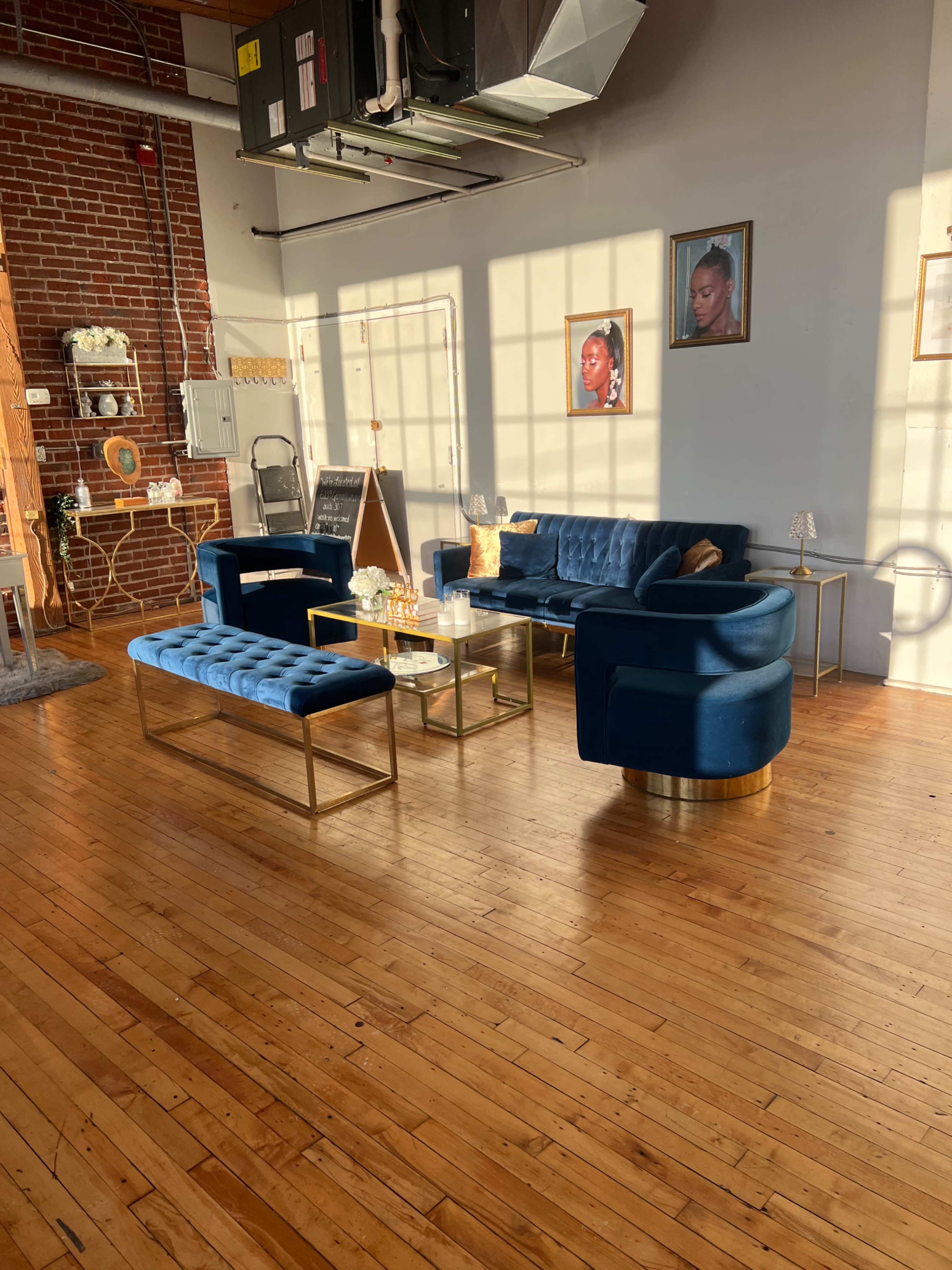 The image shows a well-lit interior space featuring a blue velvet sofa, matching benches, and framed portraits on the wall, all set against hardwood flooring.