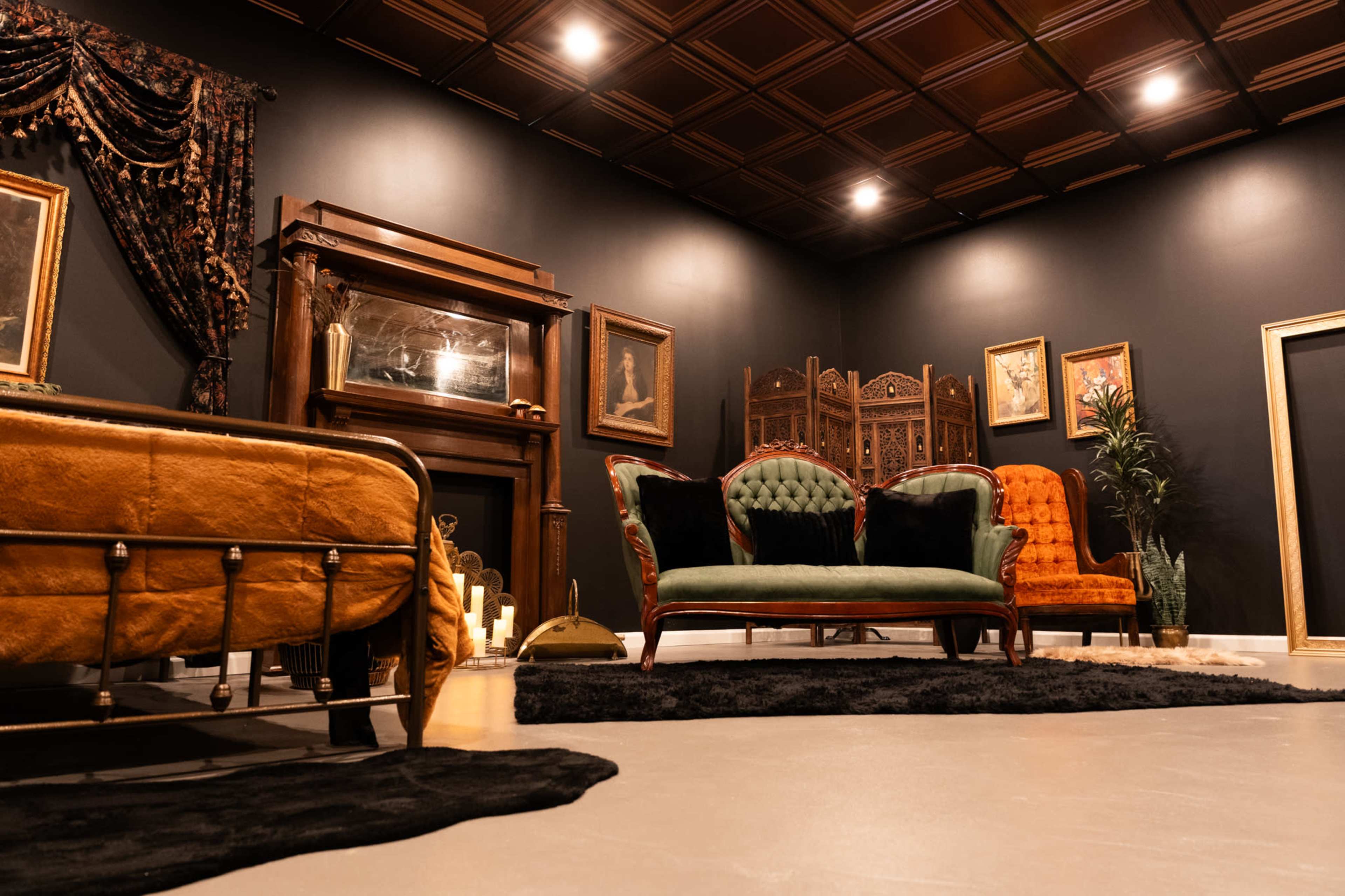 The image shows a stylish interior space featuring ornate furniture, including a green velvet loveseat, an orange armchair, and a vintage bed, all arranged around a dark backdrop with decorative elements.