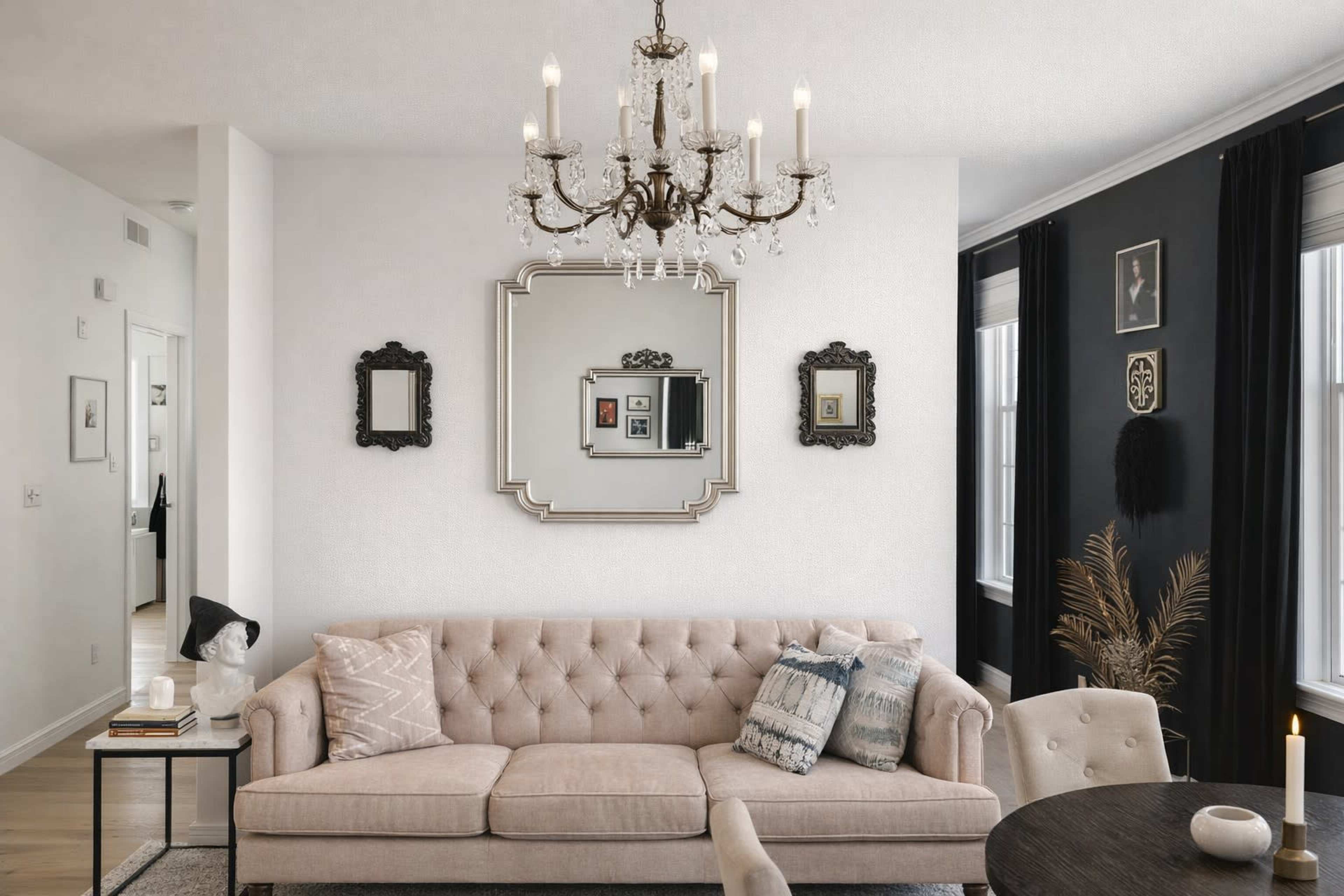 The image showcases a stylish living room featuring a beige tufted sofa, decorative mirrors on the wall, a chandelier, and a small dining table.