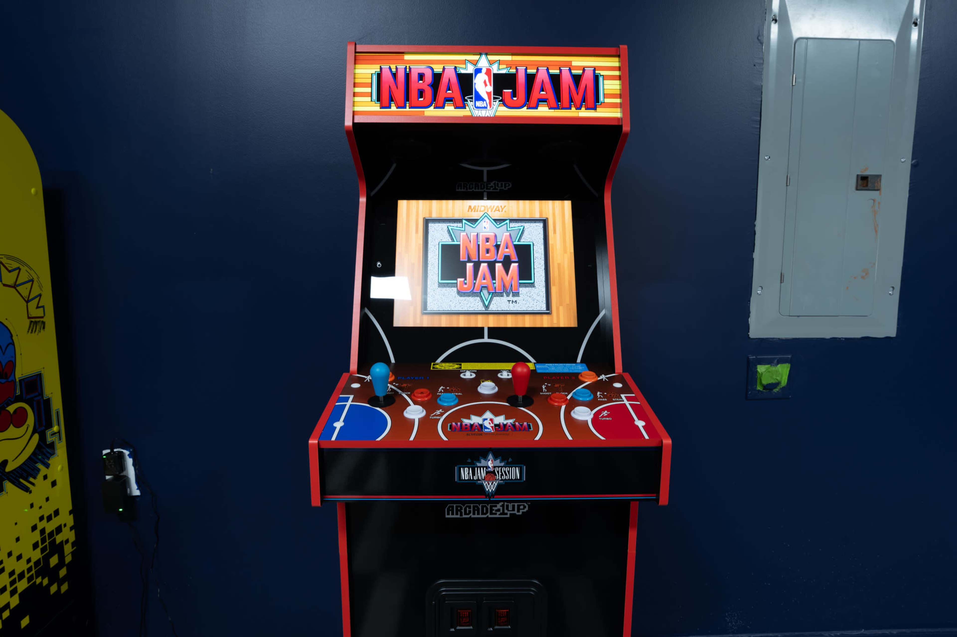 A standalone NBA Jam arcade cabinet is positioned against a blue wall, displaying the game's title on a brightly colored marquee.