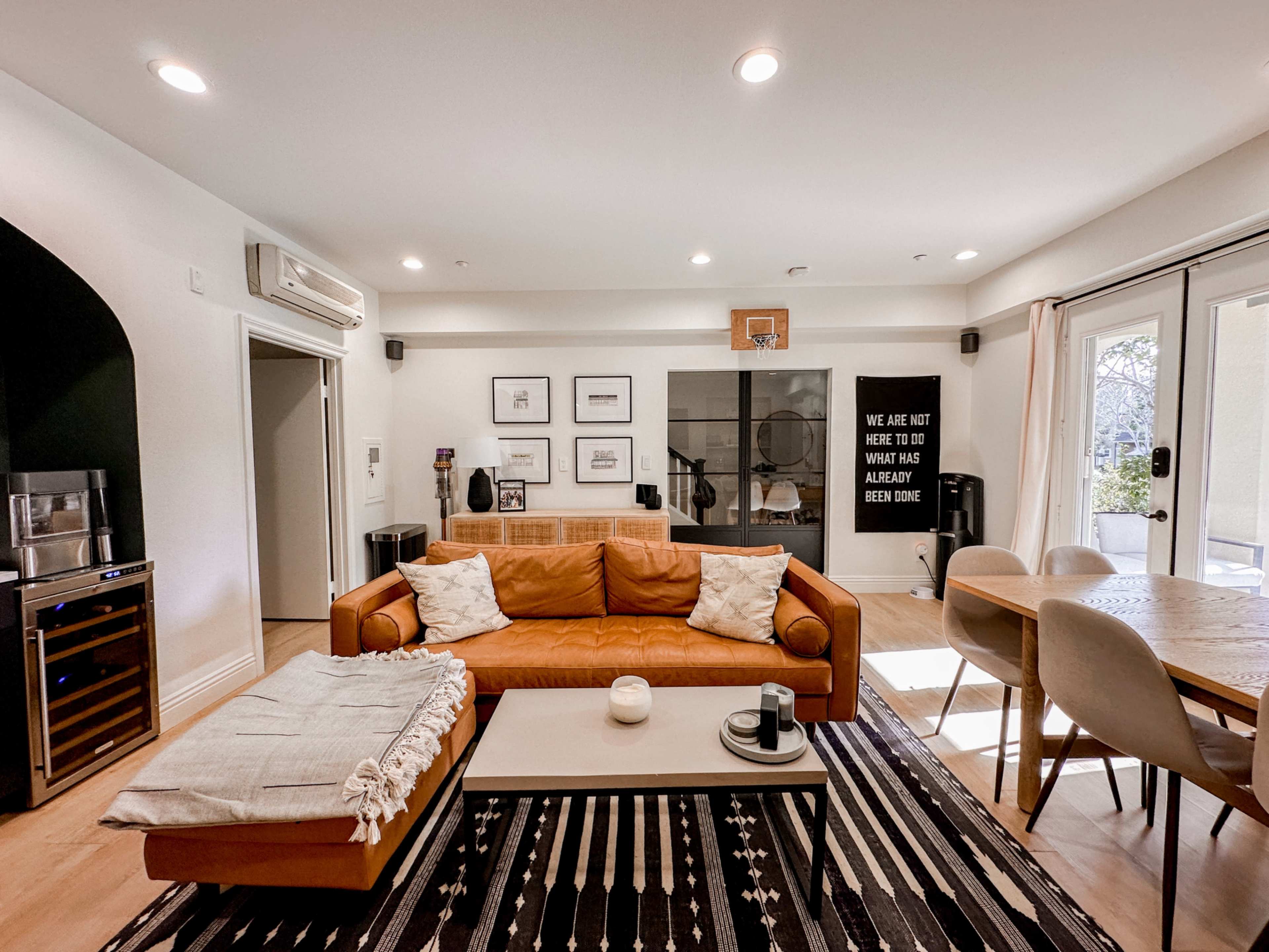 A modern living room with a brown leather sofa, a white coffee table, and a dining area next to large glass doors leading to an outdoor space.