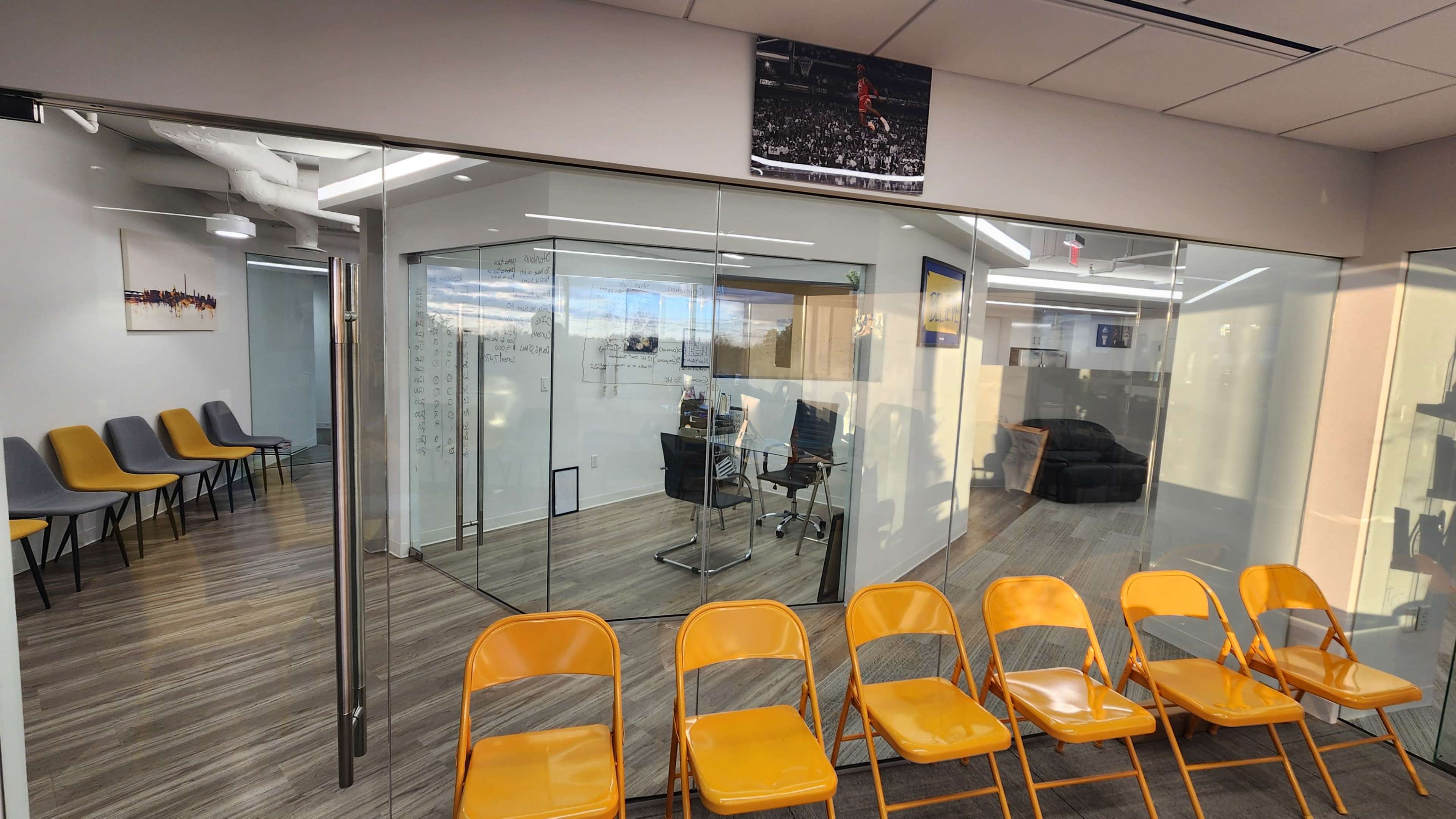 The image shows a brightly lit office space with a glass-walled meeting room and a row of orange folding chairs in the foreground.