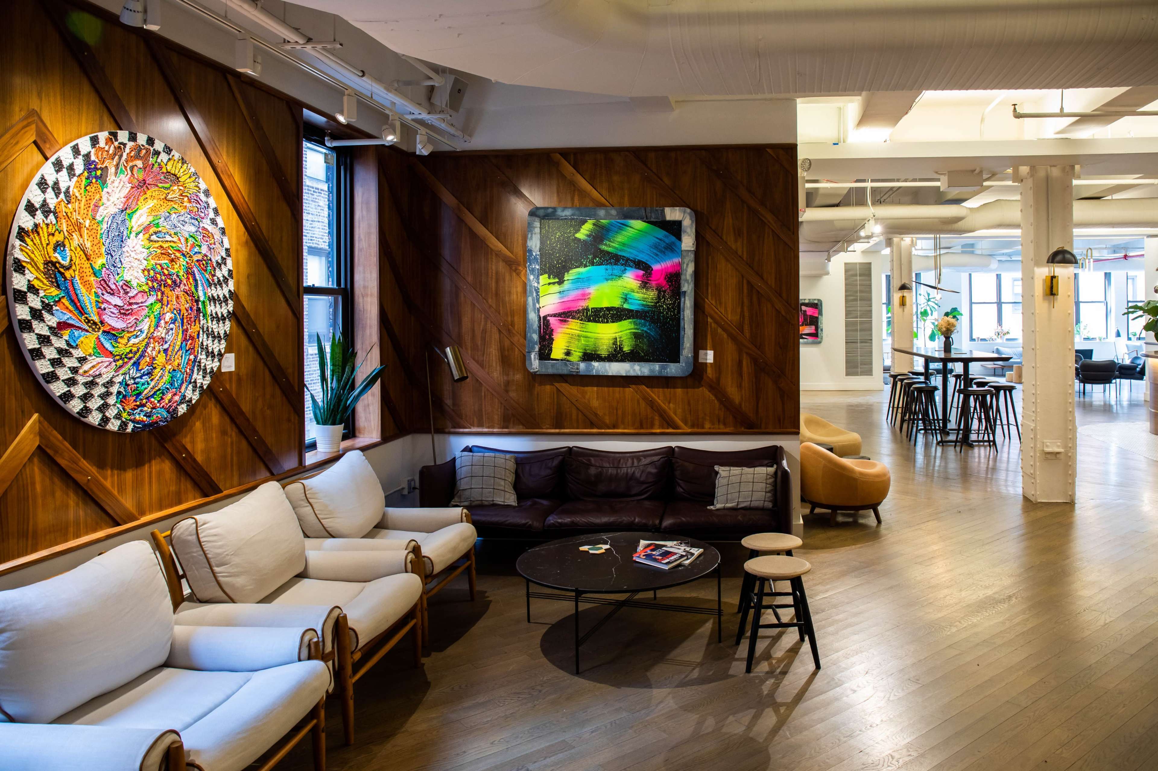 A modern lounge area featuring wooden walls, colorful artwork, a white sofa, and a round coffee table surrounded by seating.