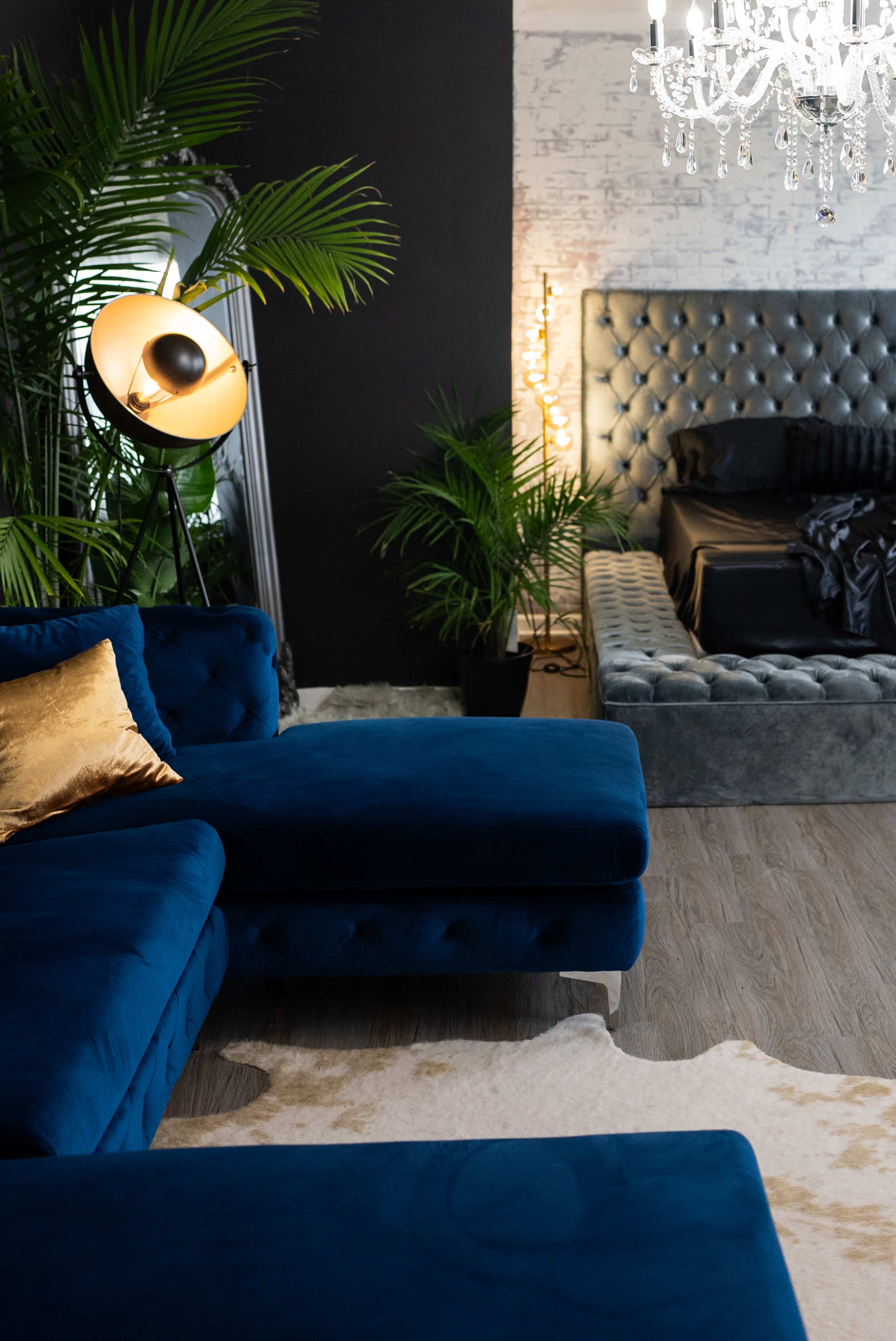 A modern living room features a blue sectional sofa, a crystal chandelier, and a tufted bed framed by tall plants.