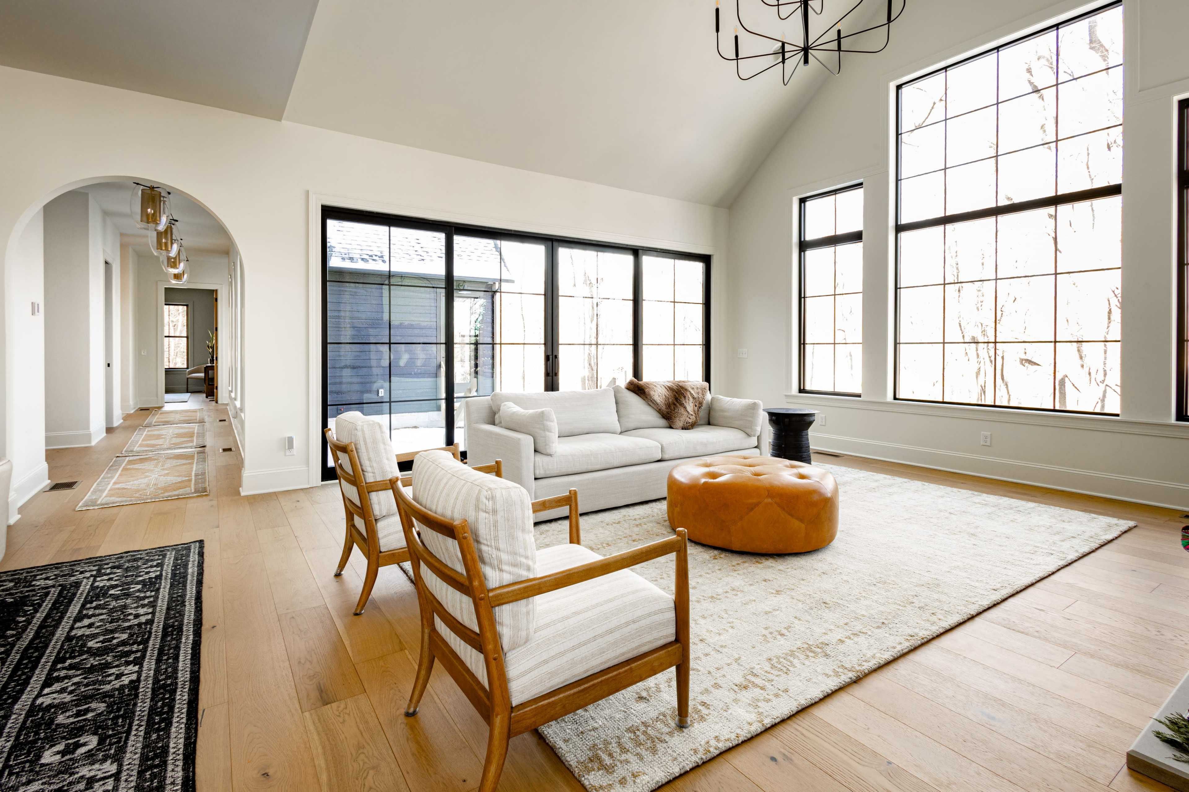 The image shows a spacious living room with large windows, a light-colored sofa, an ottoman, and wooden chairs arranged on a light hardwood floor.
