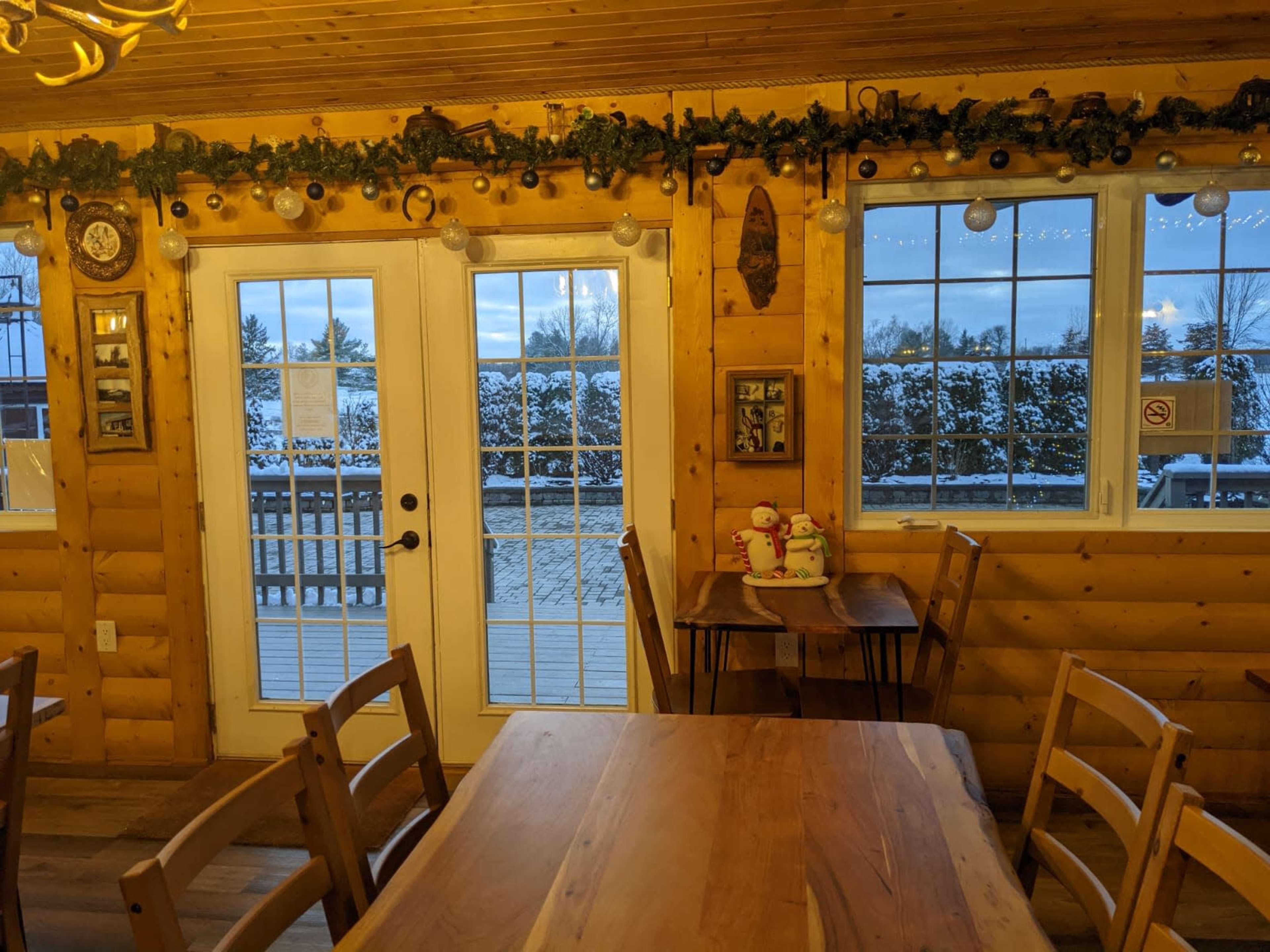A cozy wooden interior with a table and chairs, large windows displaying a snowy outdoor scene, and holiday decorations along the walls.