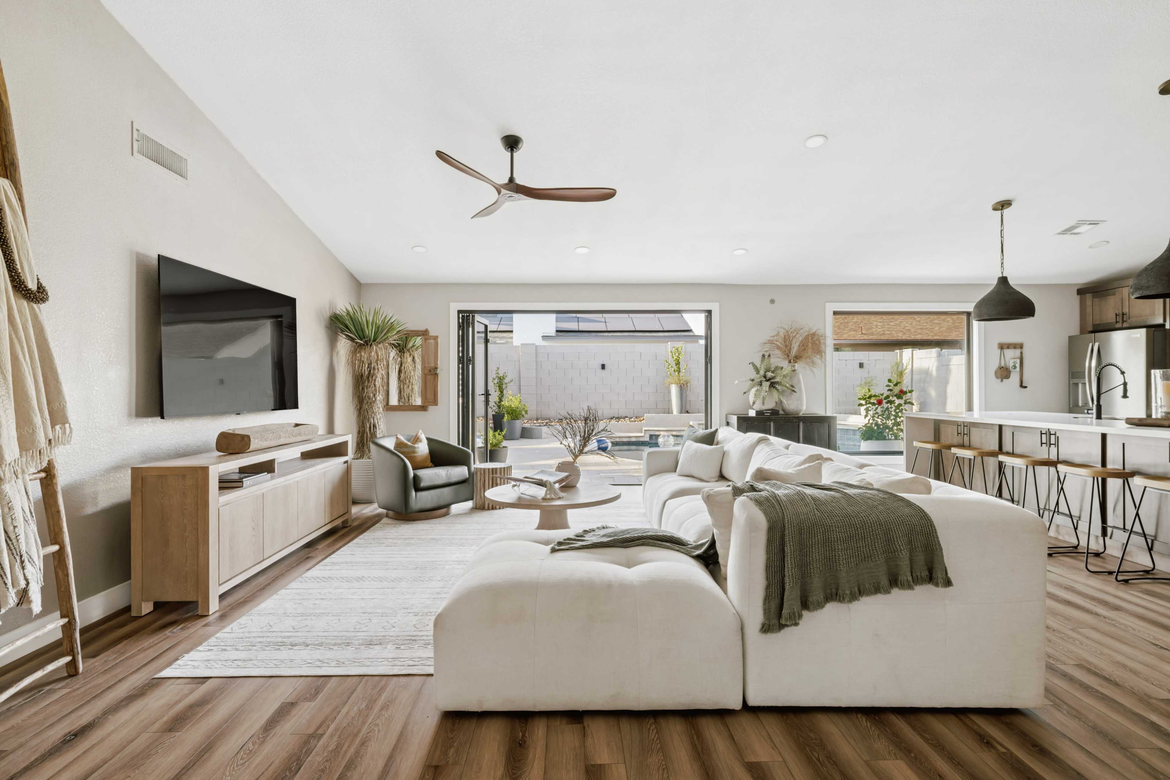 The image shows a modern living room with a large sectional sofa, a TV mounted on the wall, and sliding glass doors leading to an outdoor area.