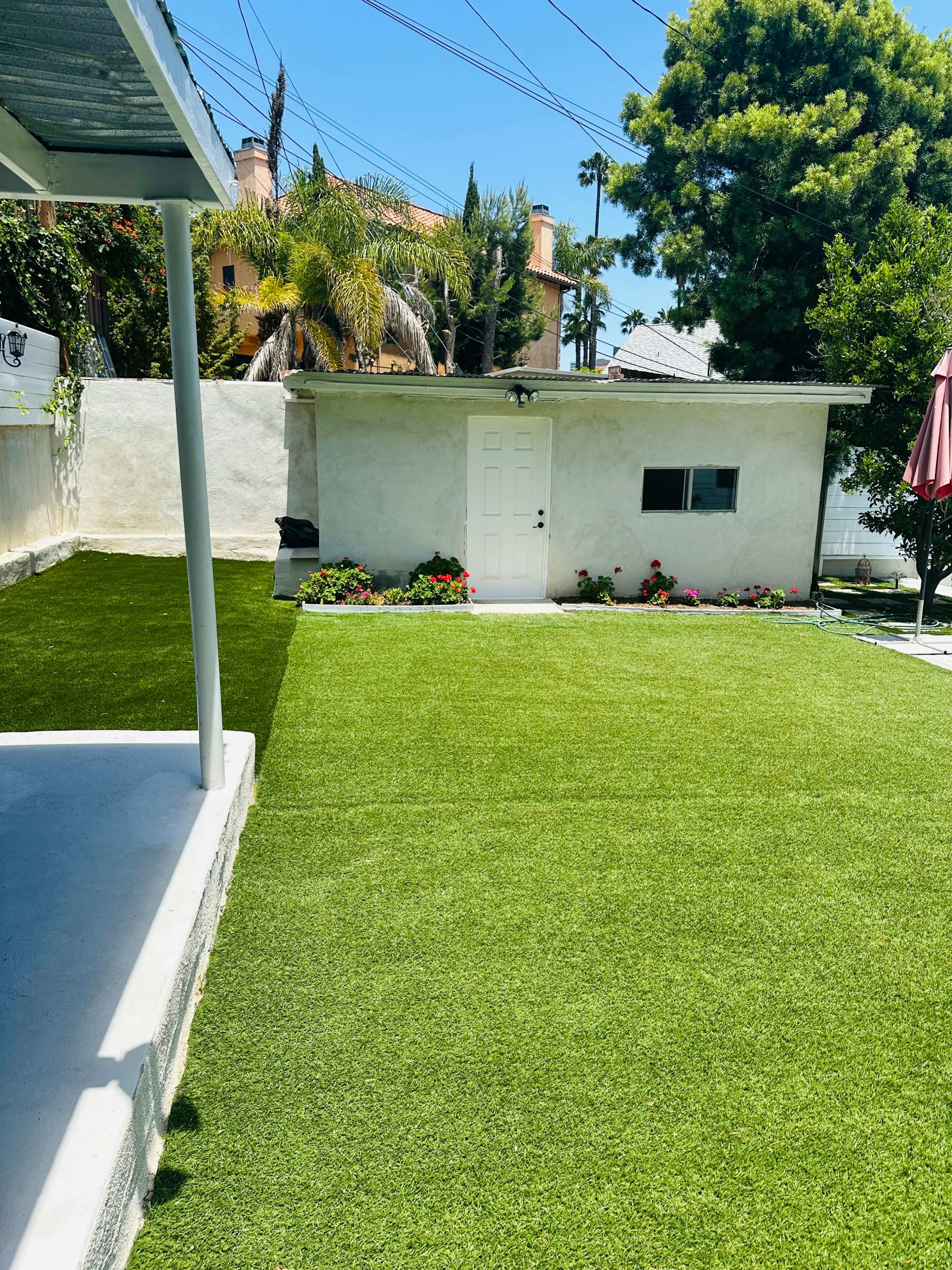 A yard with artificial grass and a small shed is visible, flanked by flowering plants and palm trees.