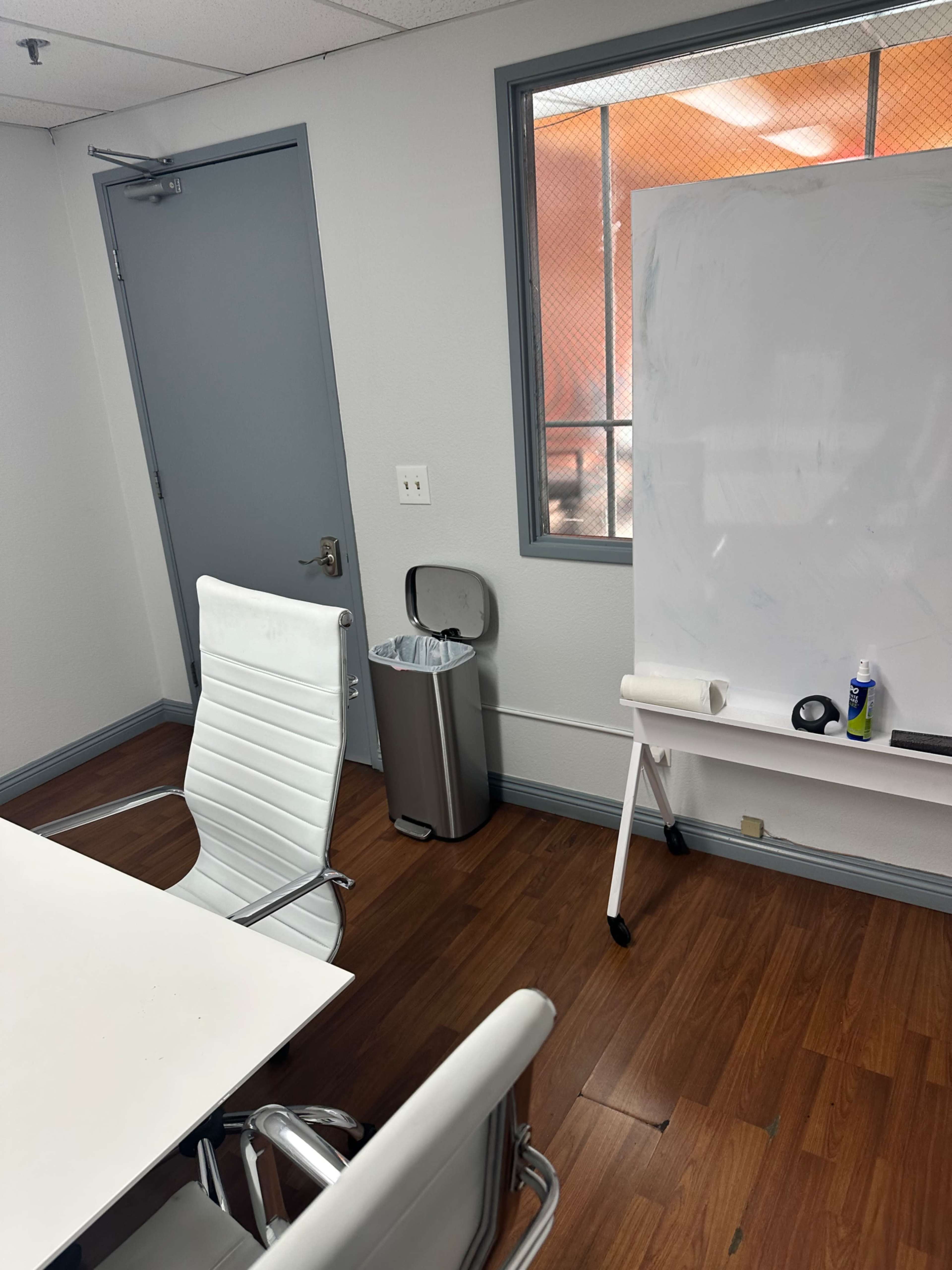 The image shows a small office space with a whiteboard, a trash can, and two white chairs around a table.