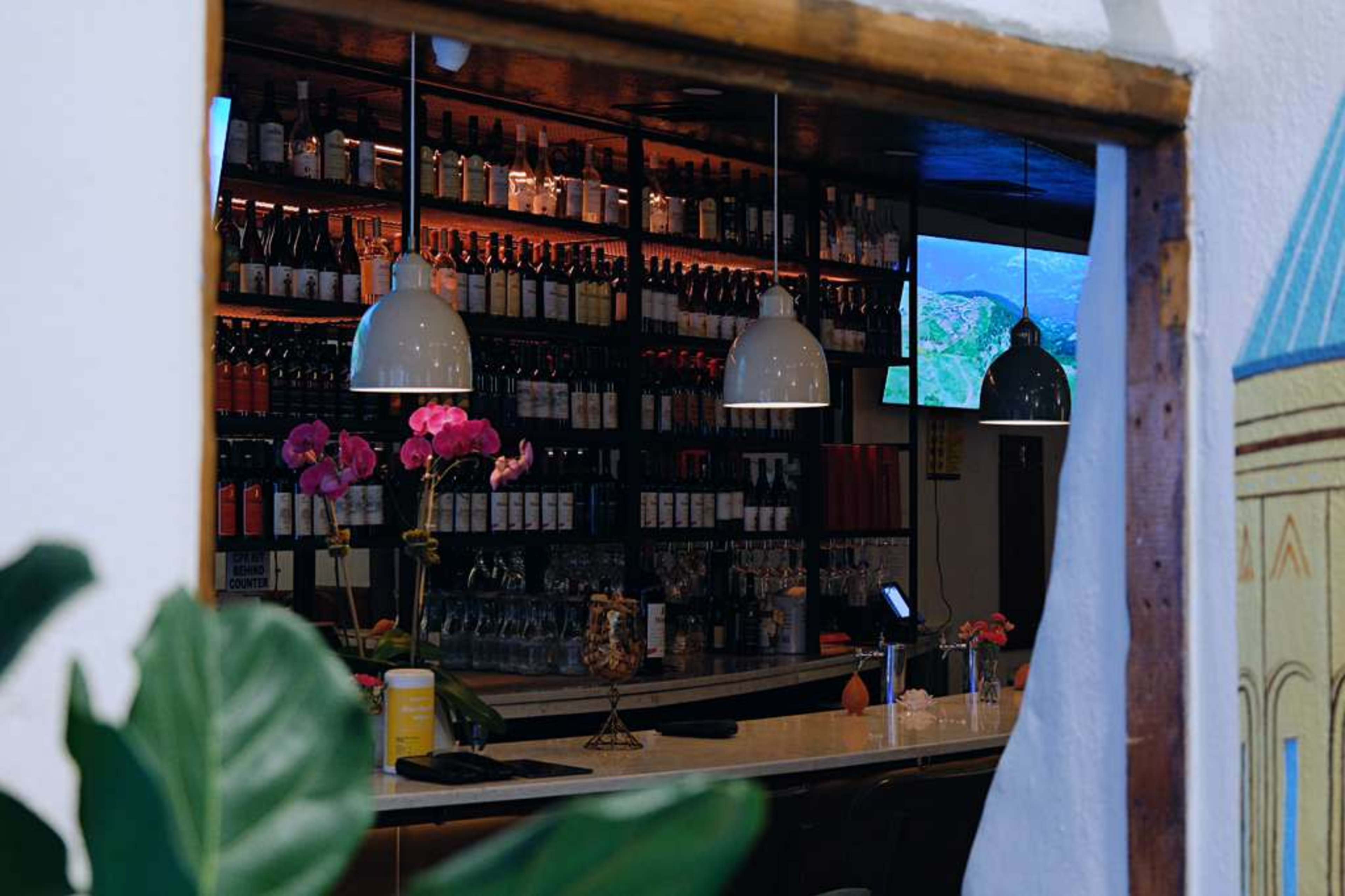 The image shows a bar area with a well-stocked shelf of bottles, two pendant lights hanging above the counter, and a television displaying a scenic view.
