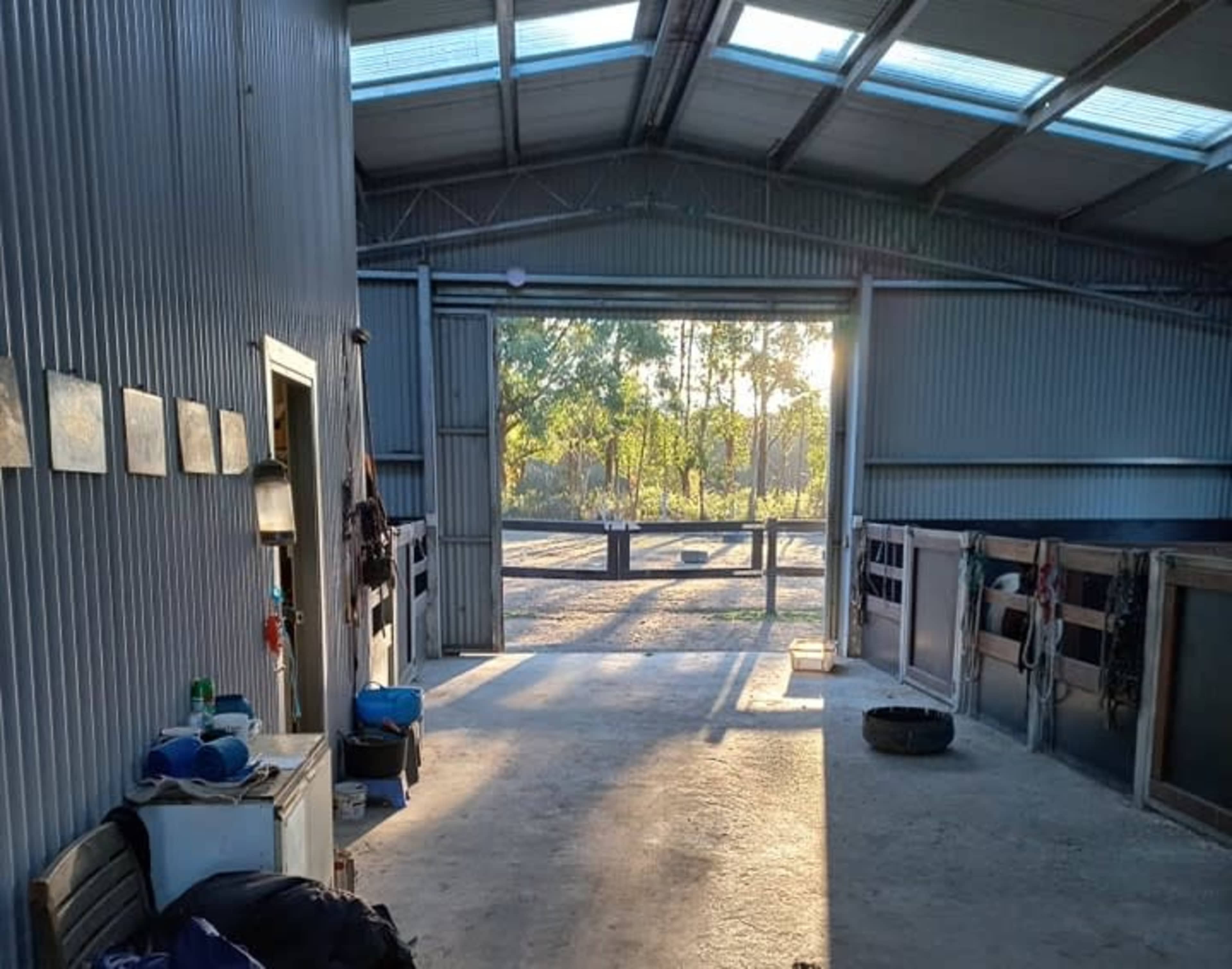 The interior of a barn shows a concrete floor, wooden stalls, and an open entrance illuminated by sunlight.
