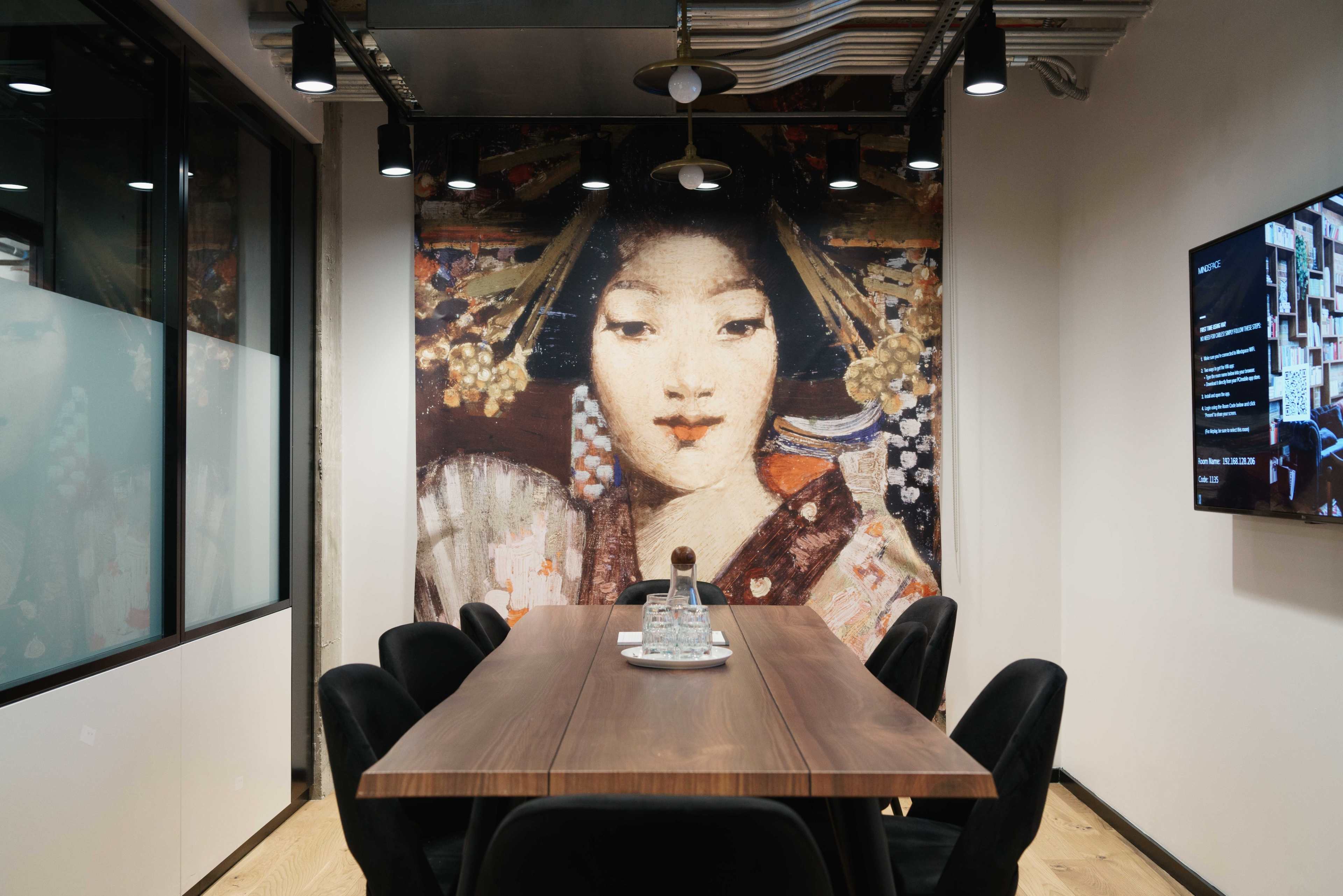 A conference room features a long wooden table surrounded by black chairs, with a large mural of a woman in traditional attire on the wall behind.