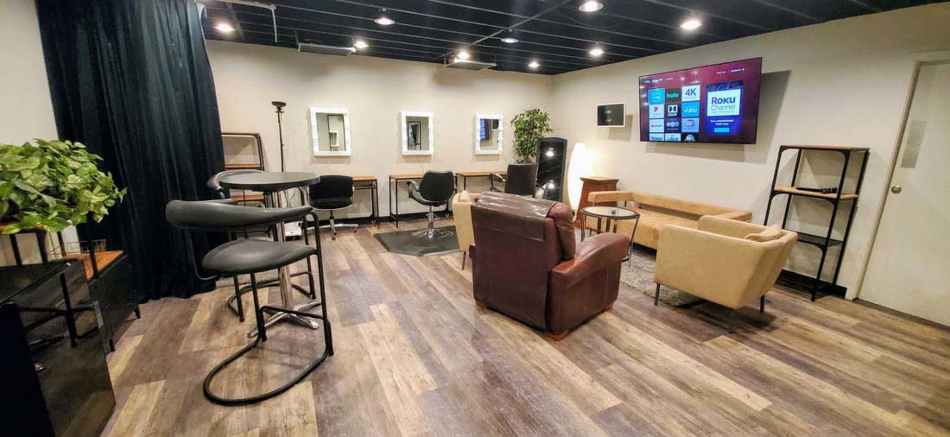 A modern lounge area features seating arrangements with a mix of chairs and tables, framed mirrors on one wall, and a large screen on another.