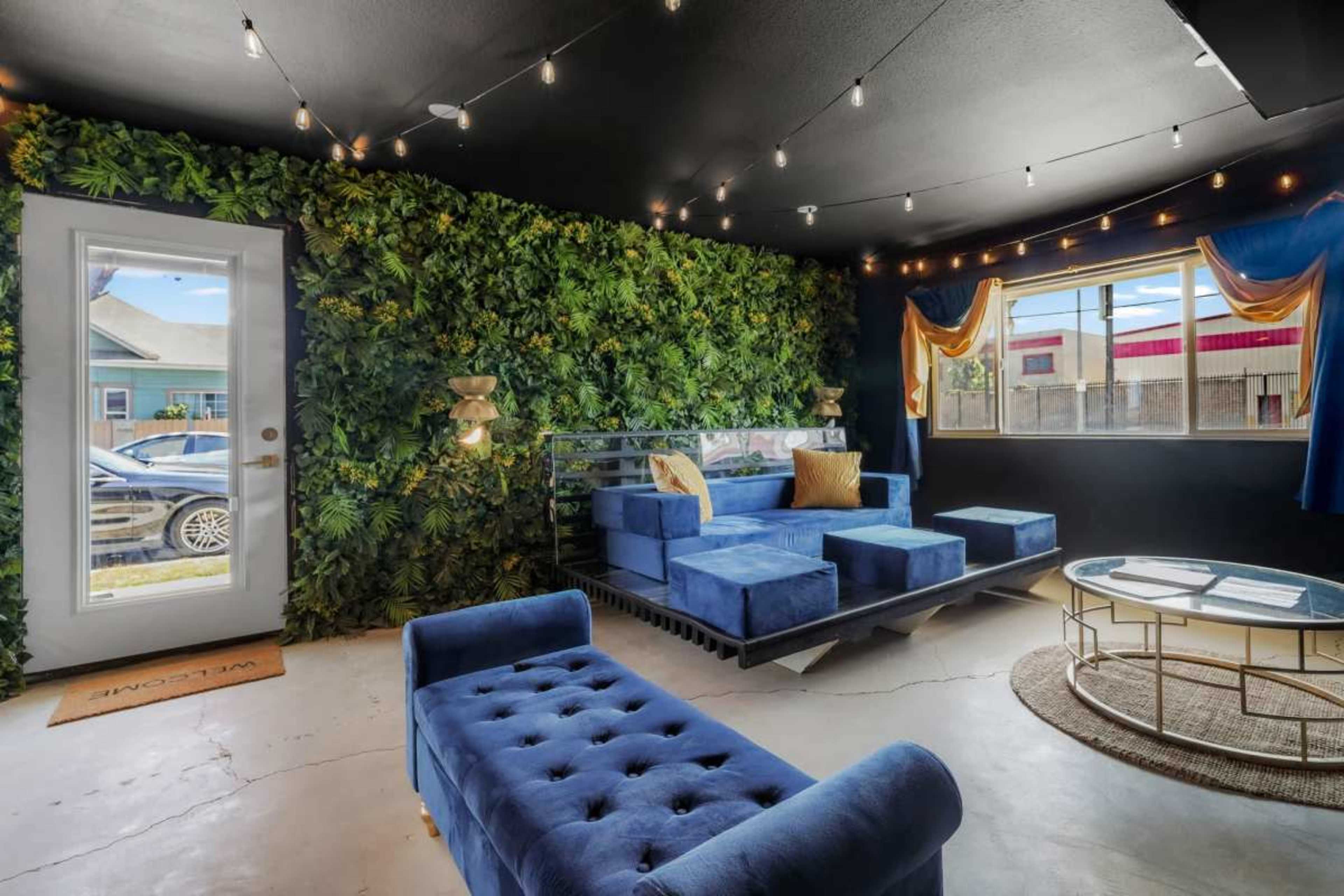 The image shows a modern room decorated with a green wall of plants, featuring a blue seating area, a circular glass table, and large windows allowing natural light to enter.