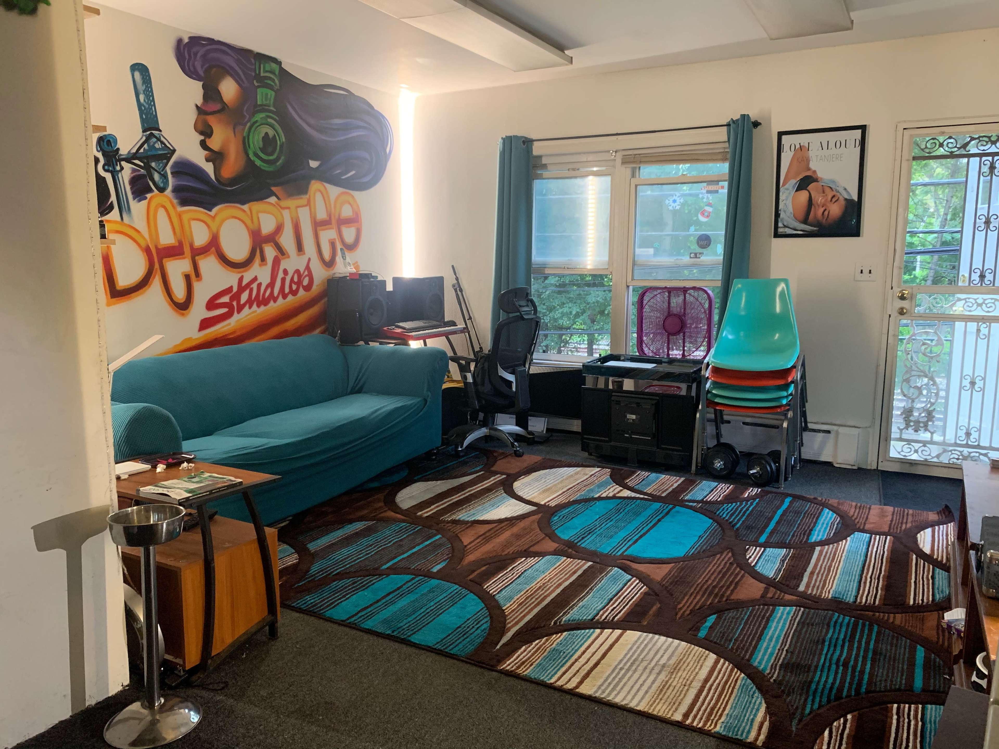 A room featuring a teal couch, a large area rug with circular designs, a mural with the text "Deportee Studios," and various furniture items including stacked chairs and a small table.