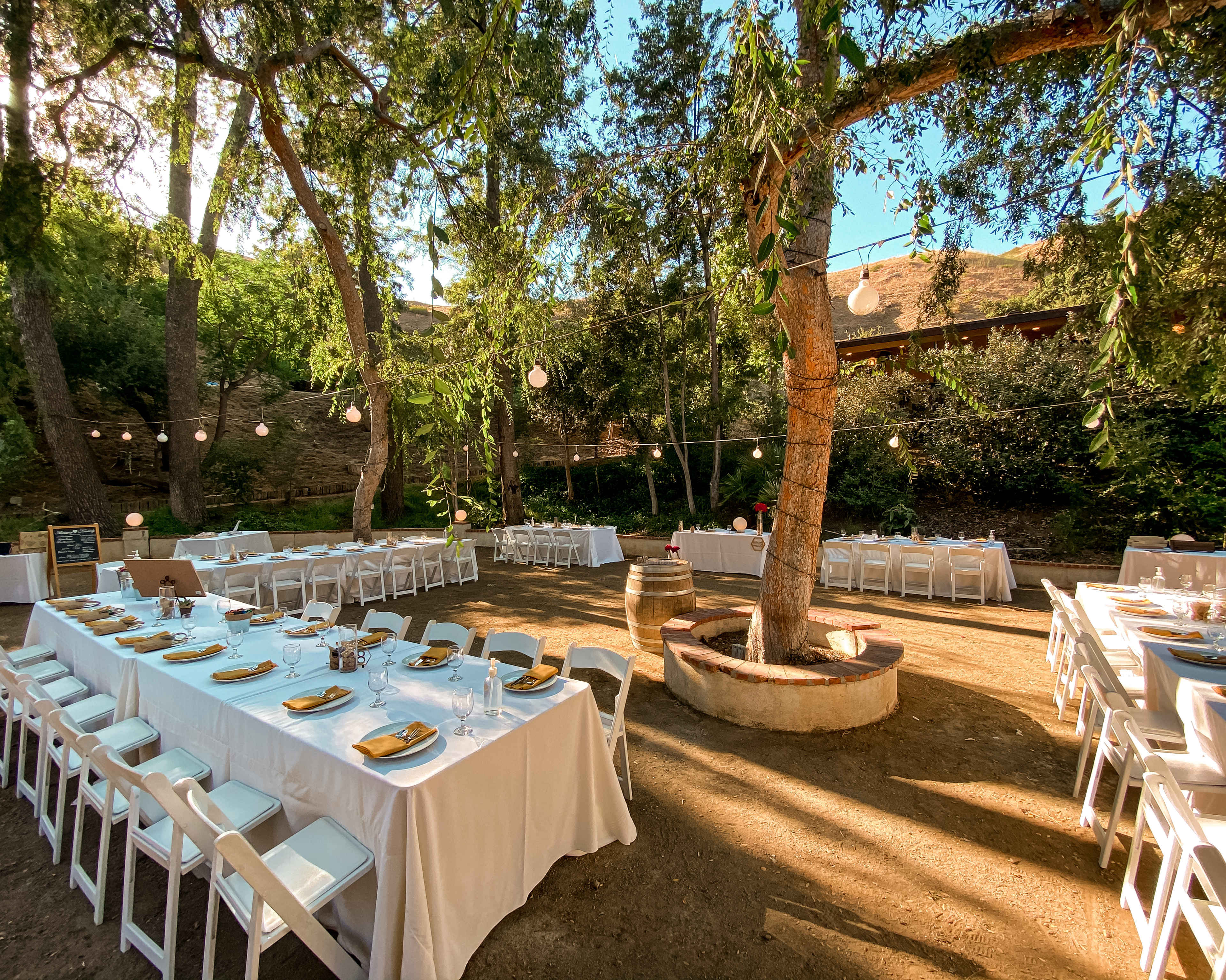 A well-arranged outdoor dining area features long tables adorned with white tablecloths, surrounded by trees and string lights.