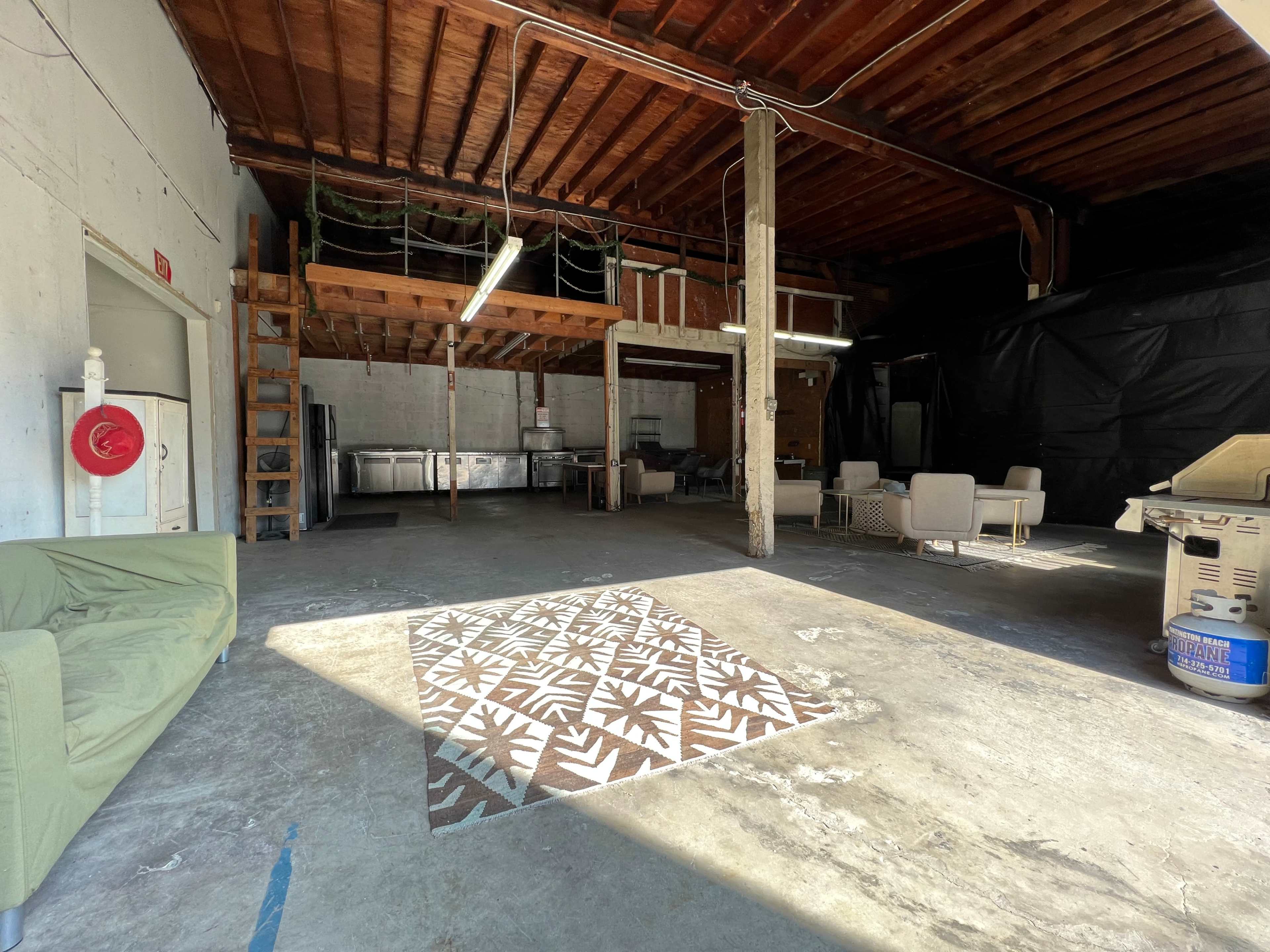 An empty industrial space with a concrete floor, exposed wooden beams, and a patterned area rug in the center.