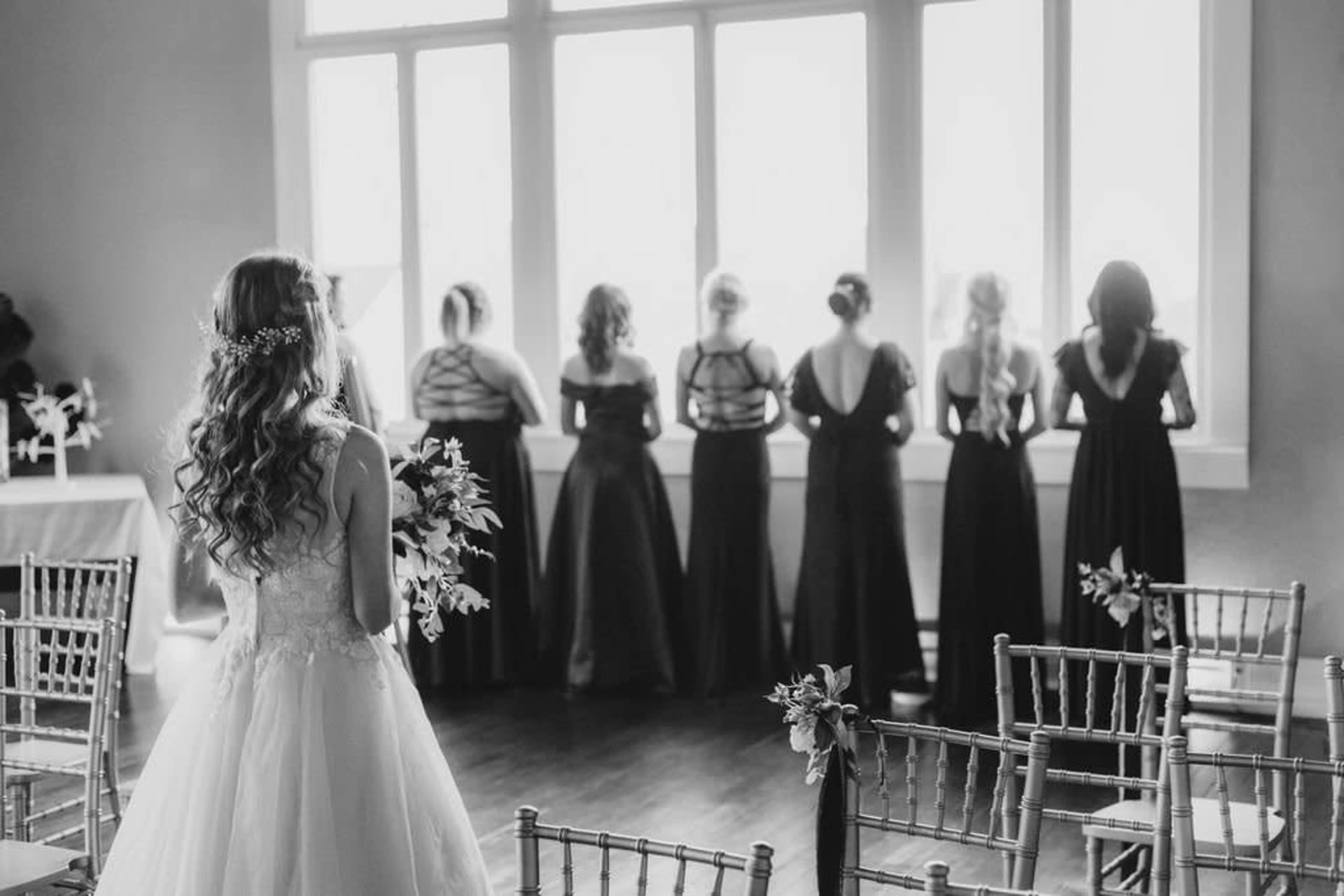 A bride in a white dress stands with her back to the camera, while her bridesmaids in black dresses gaze out the window.