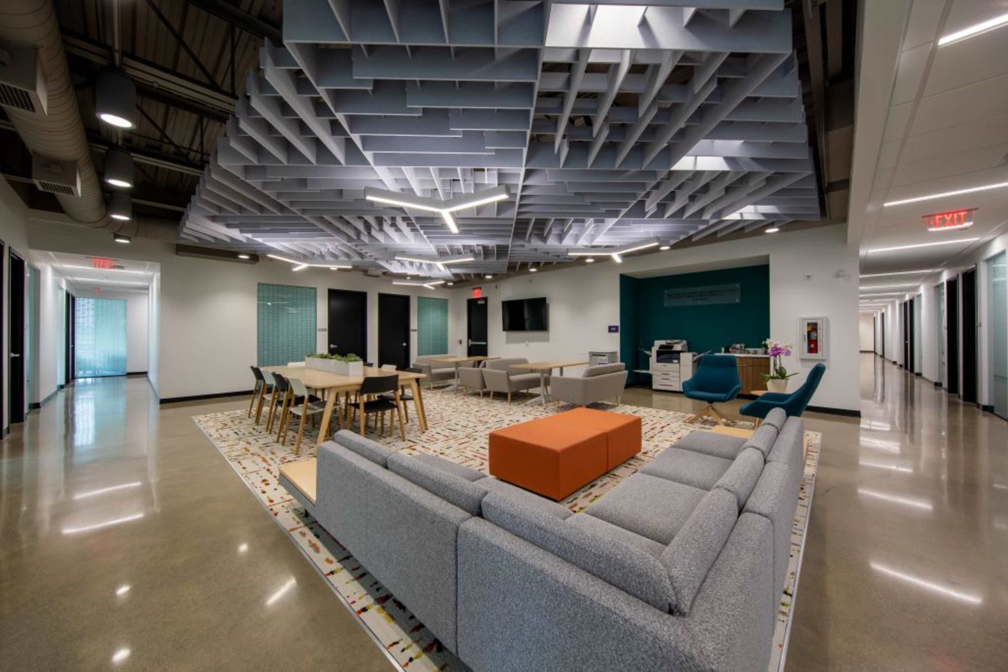 The image shows a modern office lounge area with a gray sofa, an orange ottoman, a large wooden table, and a suspended geometric ceiling design, complemented by polished concrete flooring.