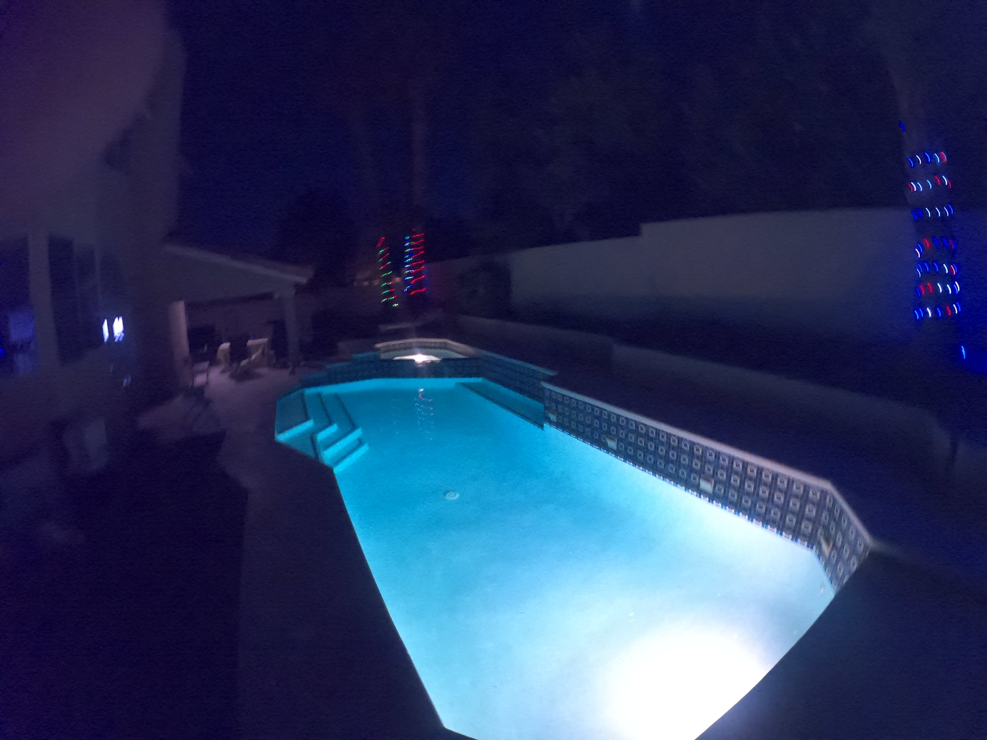 The scene features a well-lit swimming pool at night, surrounded by walls and illuminated by colorful lights.